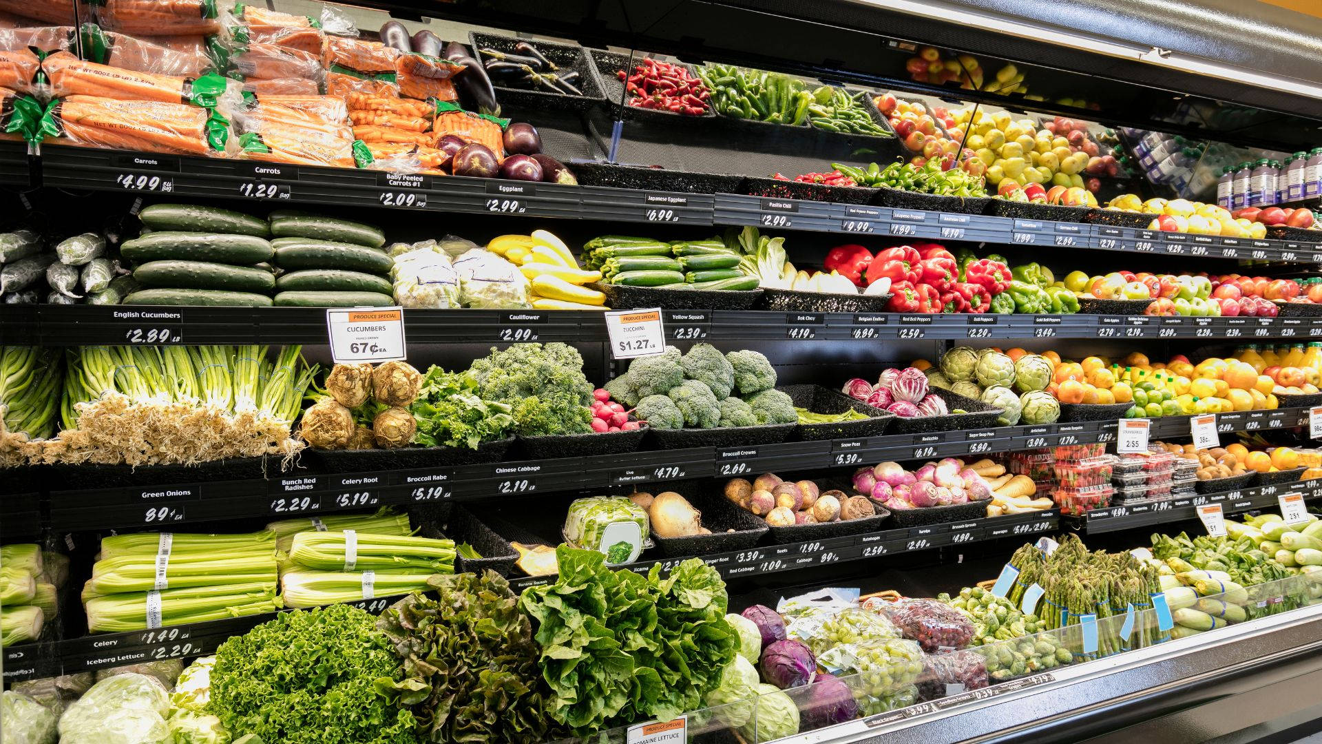 Fruits And Vegetables 桌布 壁紙 — 雜貨店內色彩繽紛的貨架上，在明亮的螢光燈下展示著各種蔬菜和水果，並附有價格標籤。