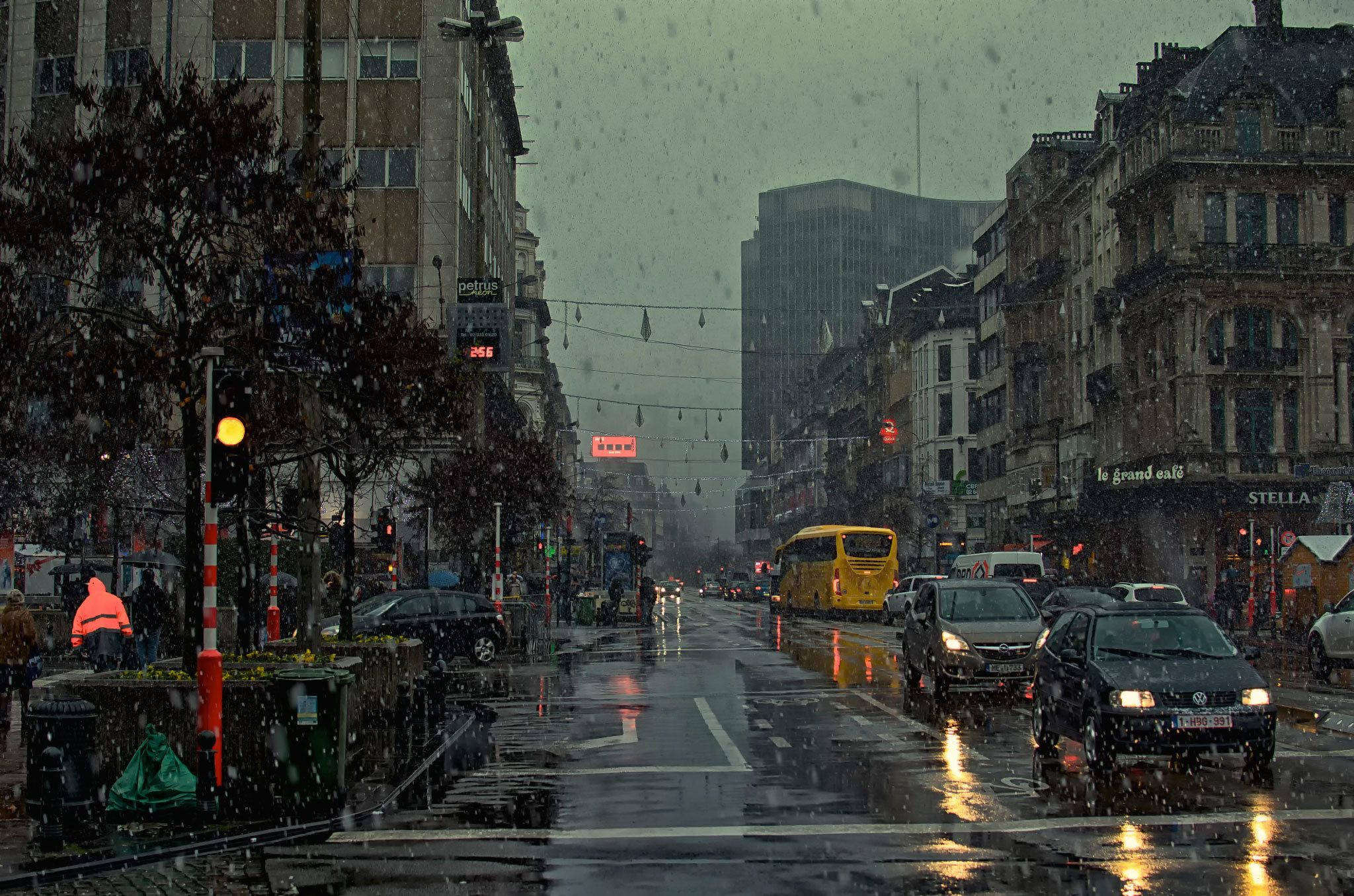 下雨 桌布 壁紙 — 雨中的城市街道場景，濕漉漉的人行道反射著車燈，一輛黃色巴士在飄落的雪中行駛。