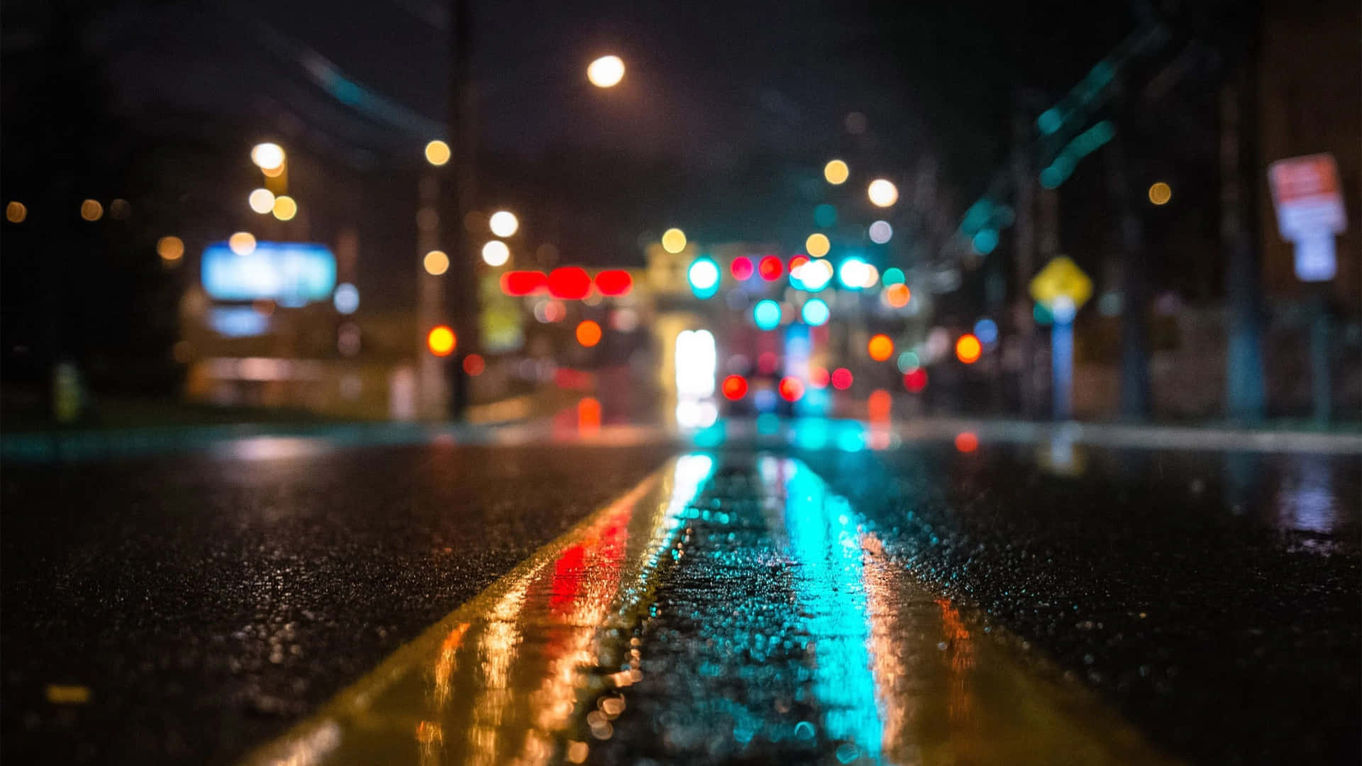 下雨 桌布 壁紙 — 雨夜中濕滑的都市街道，發光的散景燈光映照在濕瀝青和黃色車道標記上。