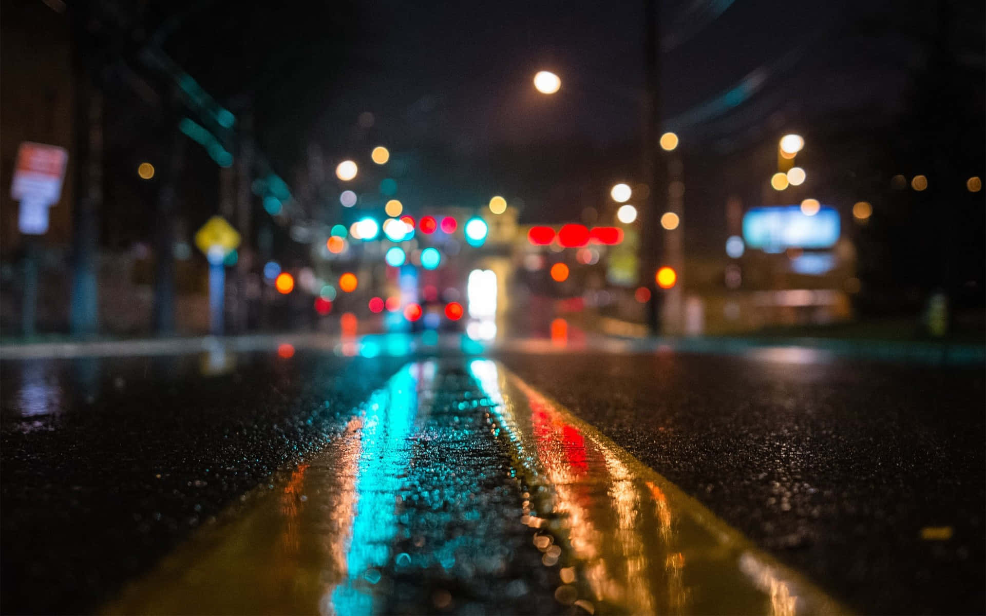 桌布 壁紙 — 雨夜中濕漉漉的城市街道，發光的散景燈光反射在潮濕的瀝青和黃色車道標記上。
