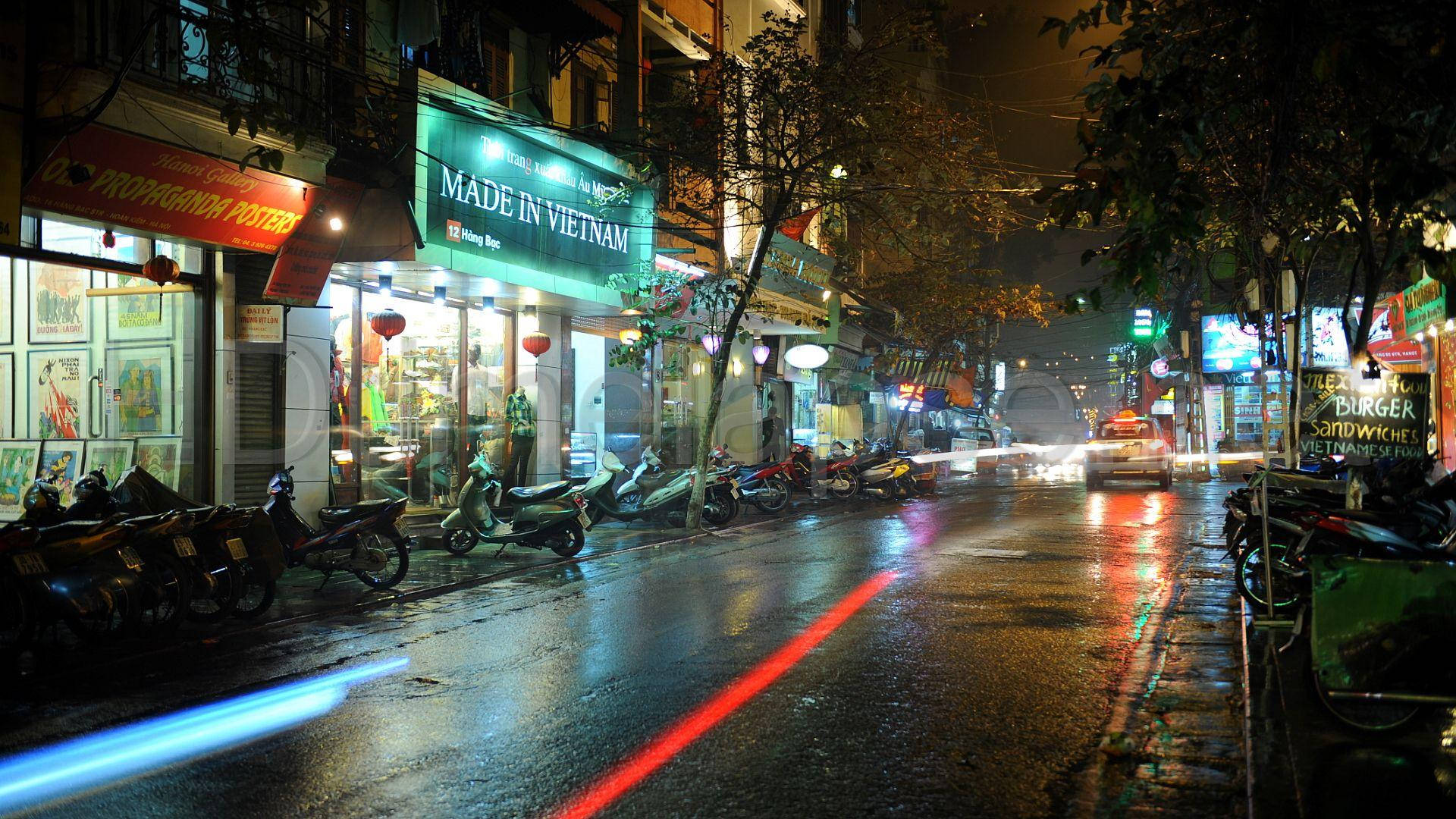 Hanoi 桌布 壁紙 — 雨夜中濕漉漉的城市街道，霓虹招牌和汽車燈光在濕漉漉的路面上反射出光芒。