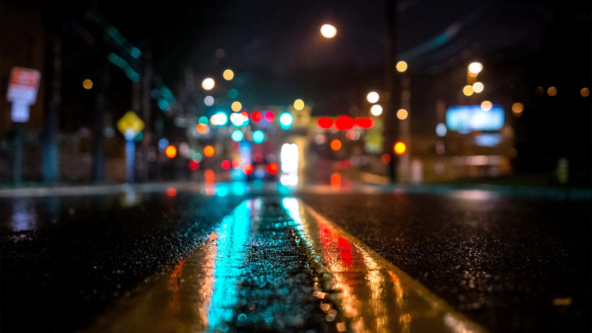 黑暗城市 桌布 壁紙 — 雨夜中的城市街道，濕漉漉的瀝青路面和黃色車道標線反射著閃爍的散景燈光。