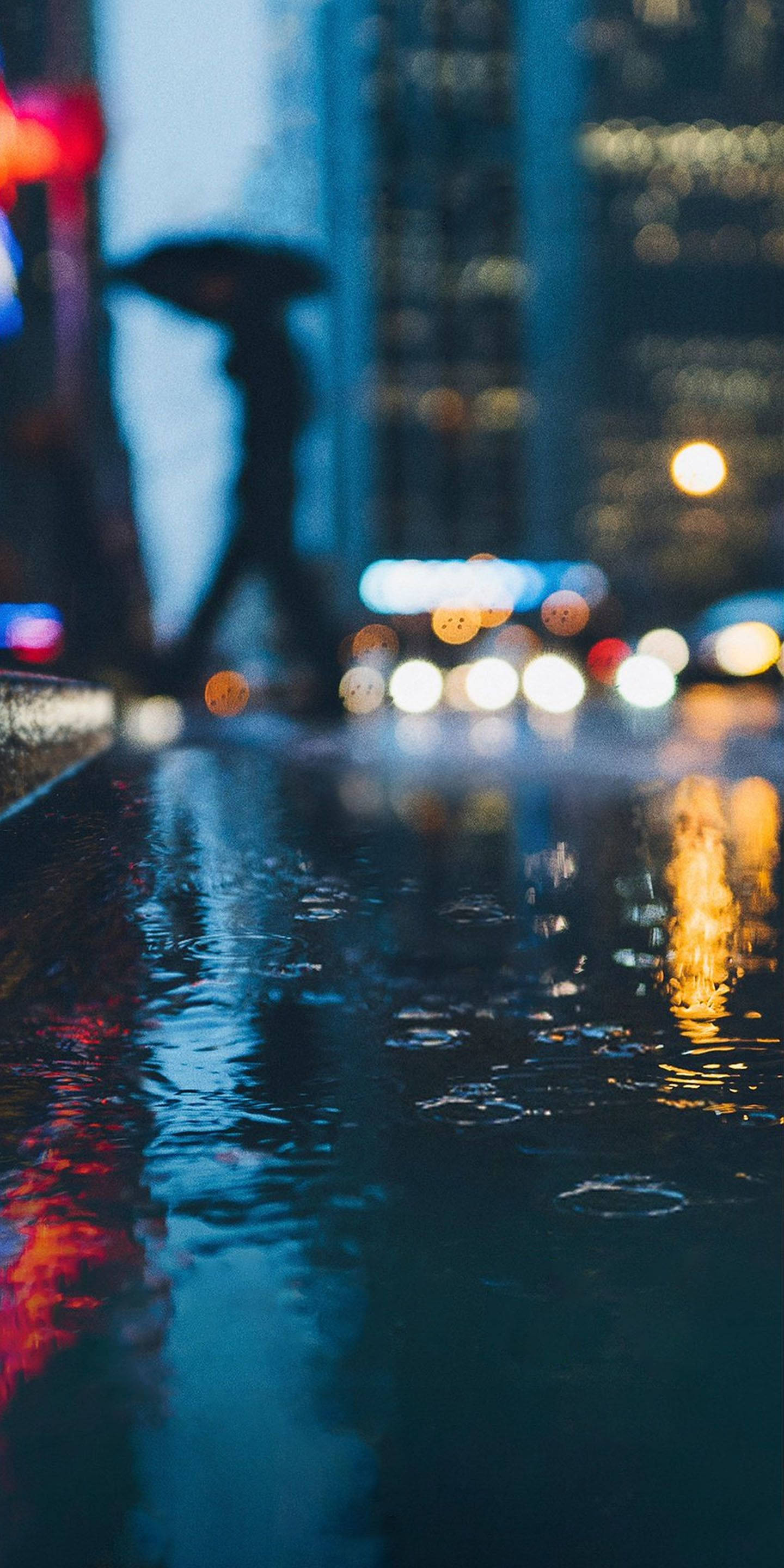 渡我被身子 桌布 壁紙 — 雨夜城市景觀，模糊的燈光在濕漉漉的人行道上反射，一個孤獨的身影撐著雨傘。