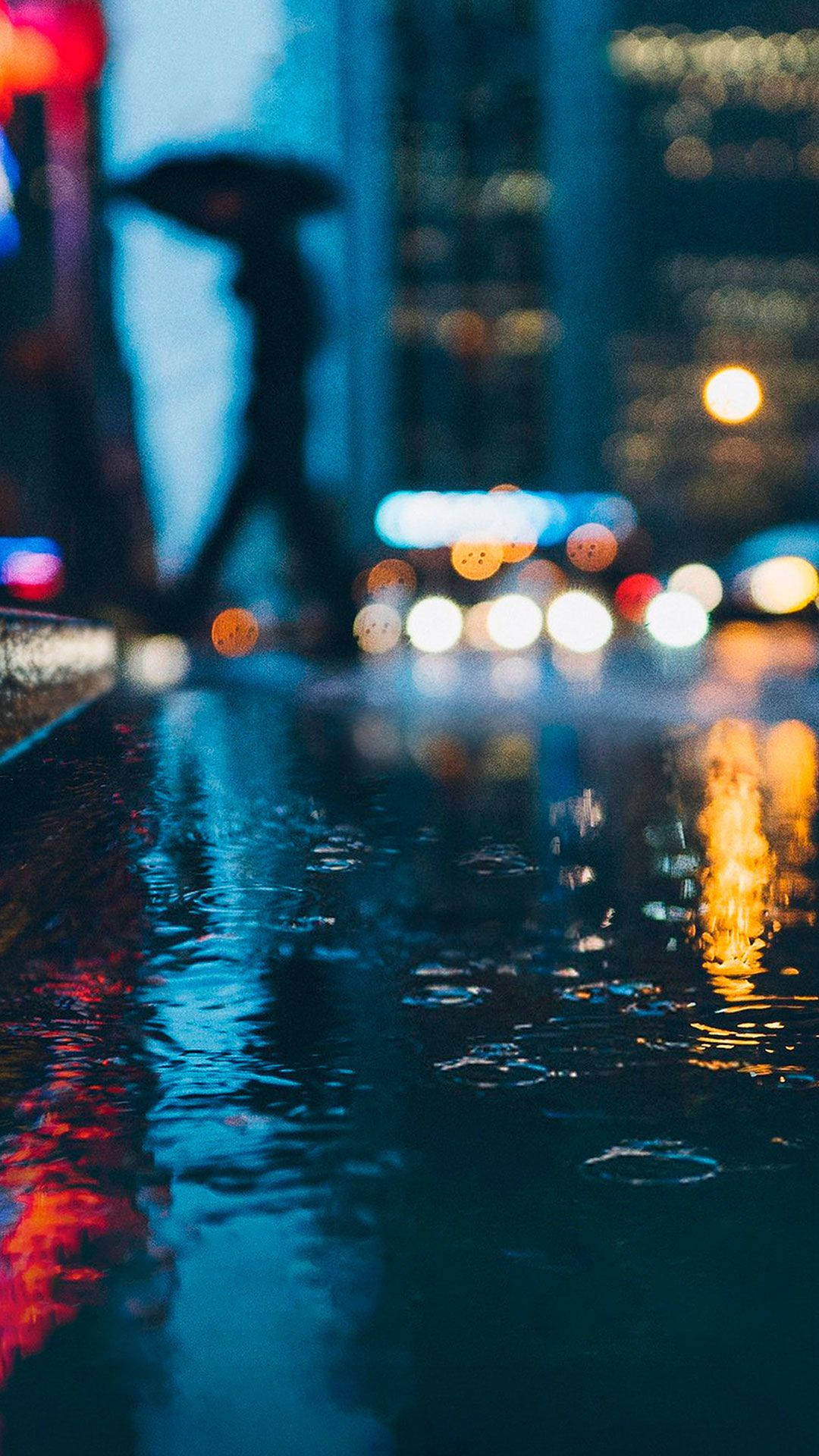 Street 桌布 壁紙 — 雨夜城市景觀，模糊的燈光在濕漉漉的人行道上反射，一個撐著雨傘的剪影人物。