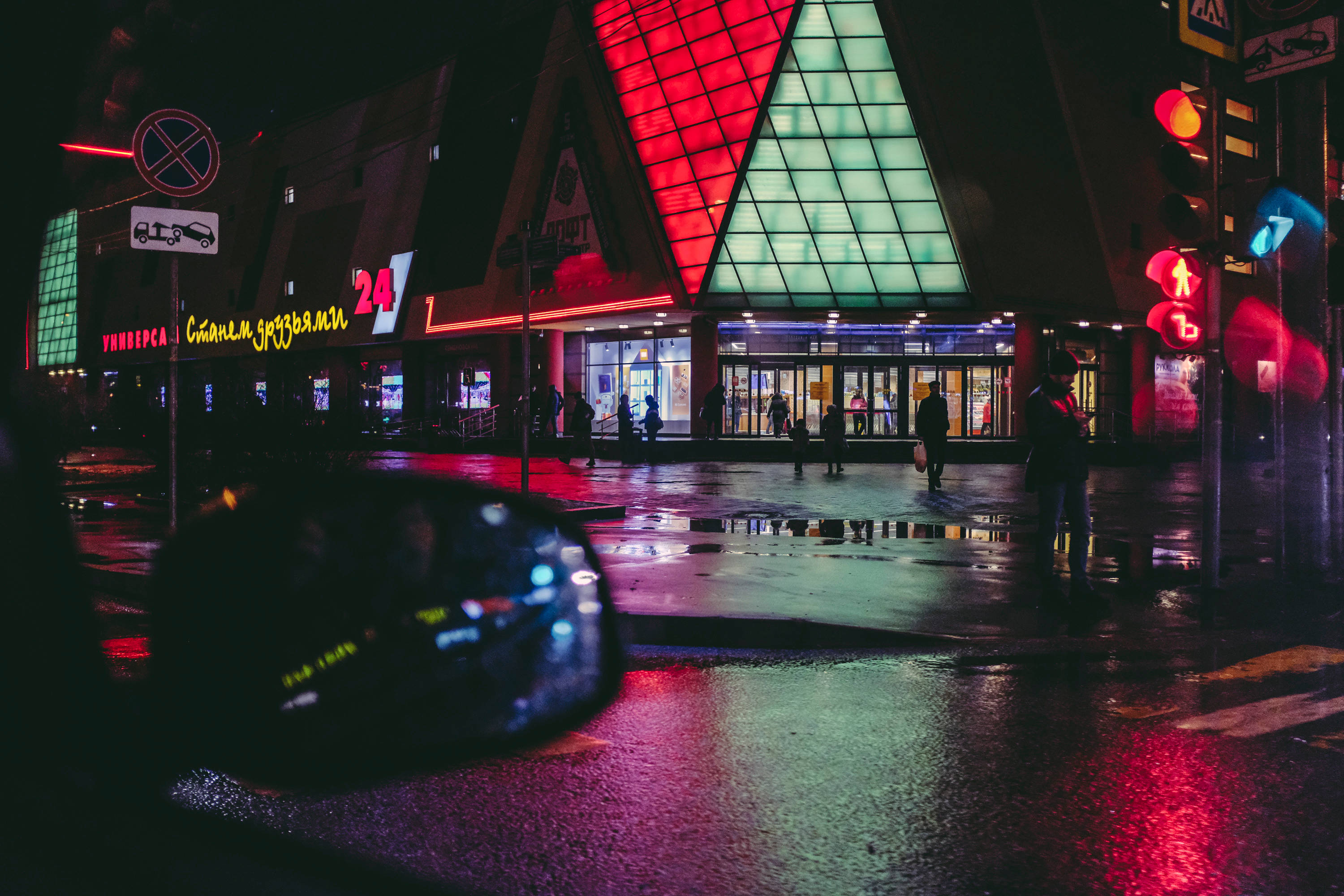 霓虹美學 桌布 壁紙 — 雨夜城市景觀，霓虹燈光在濕漉漉的人行道上反射，行人靠近一座明亮照明的三角形建築。