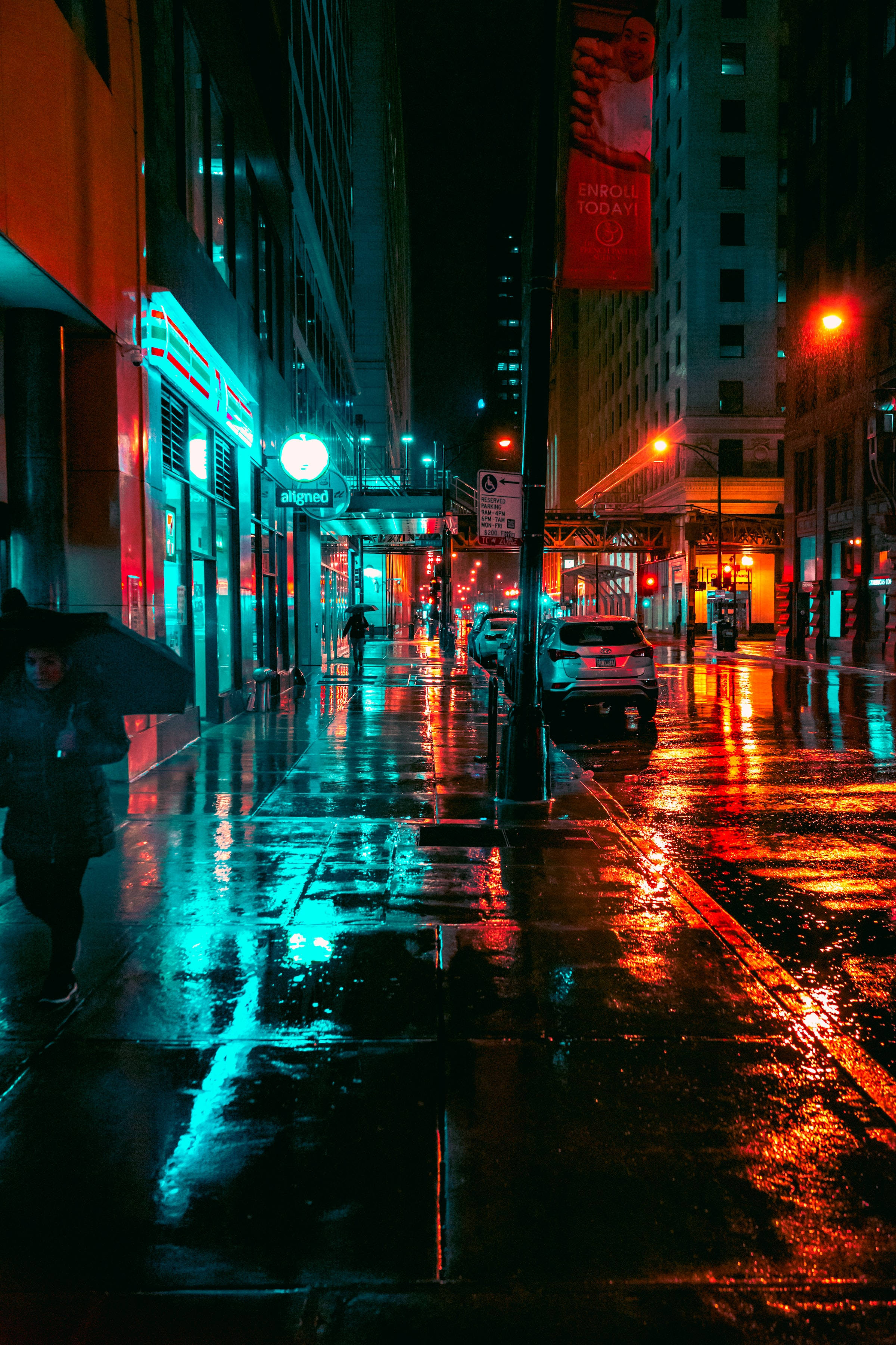 夜空 桌布 壁紙 — 雨夜霓虹燈照亮的城市街道，濕漉漉的人行道反射著商店櫥窗和路燈的鮮豔紅色與青色光芒。