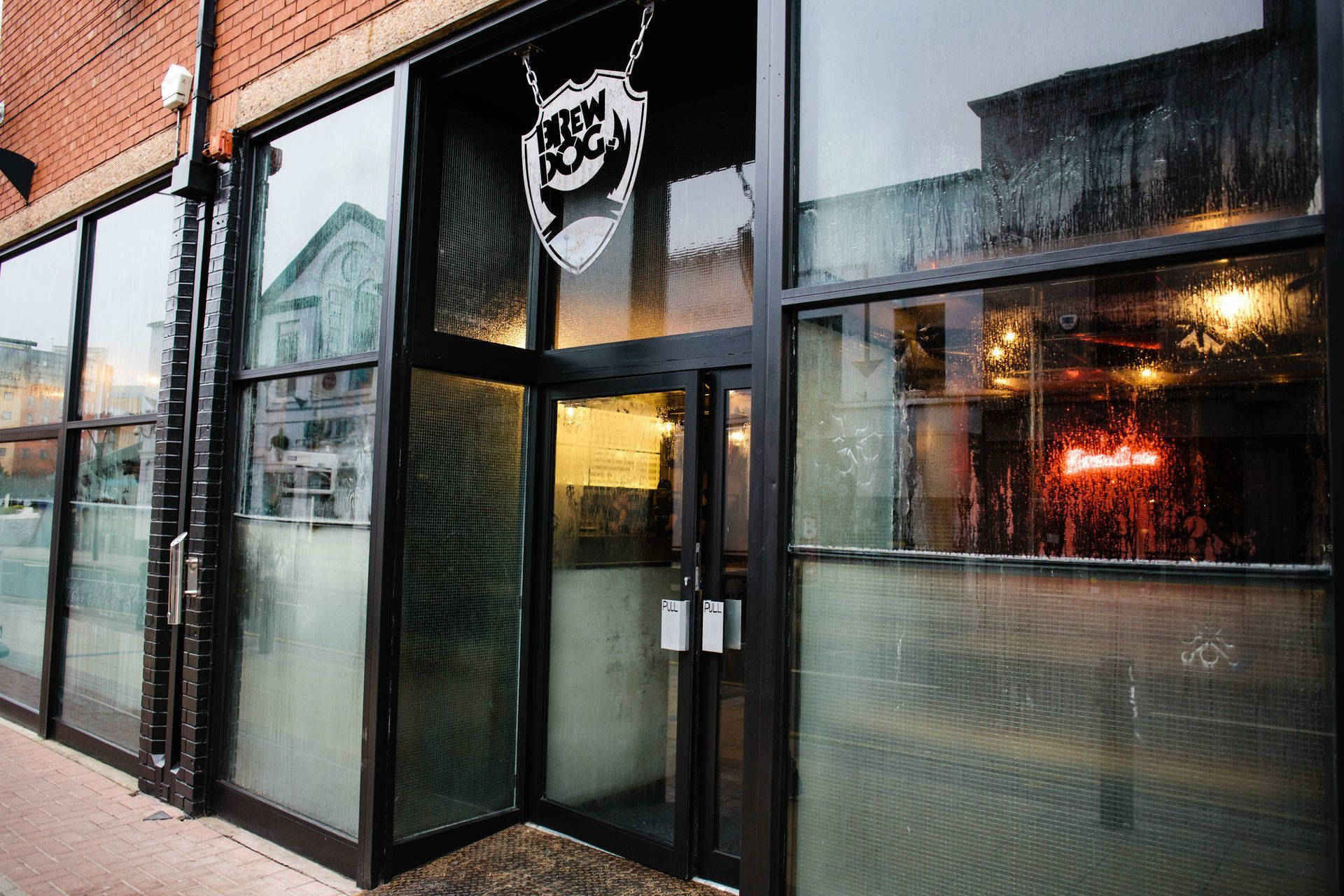 Drink 桌布 壁紙 — 雨天的BrewDog酒吧入口景觀，濕漉漉的玻璃窗反射城市燈光，內部有發光的霓虹燈牌。