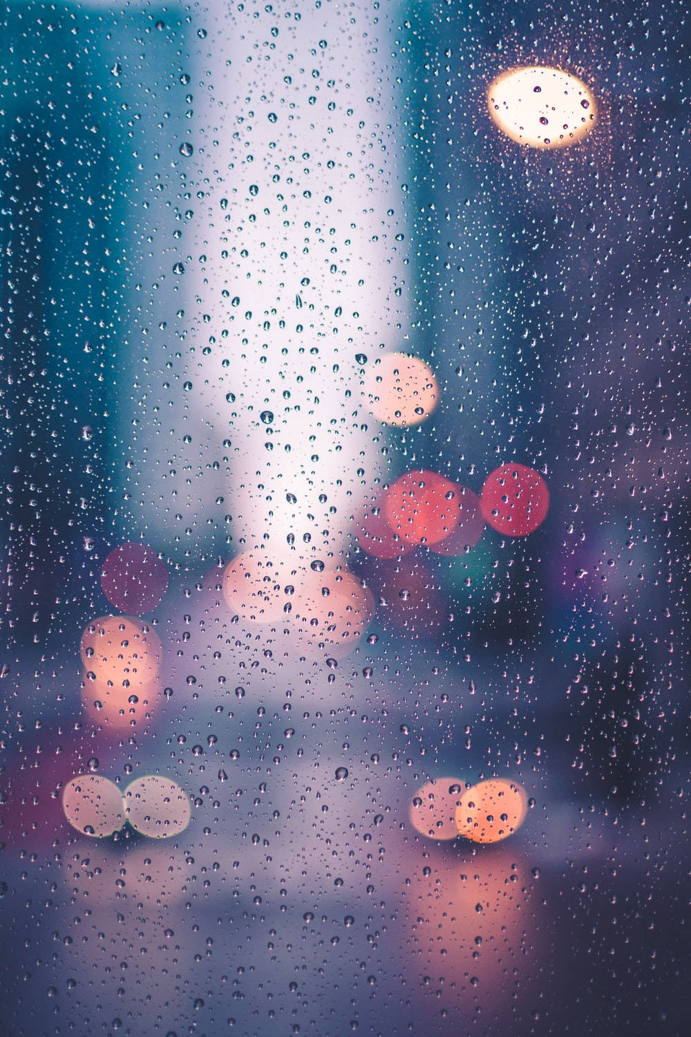 Rainy Day 桌布 壁紙 — 雨滴依附在窗戶上，顯露出模糊的城市燈光，在清涼的藍色暮色背景下溫暖地發光。