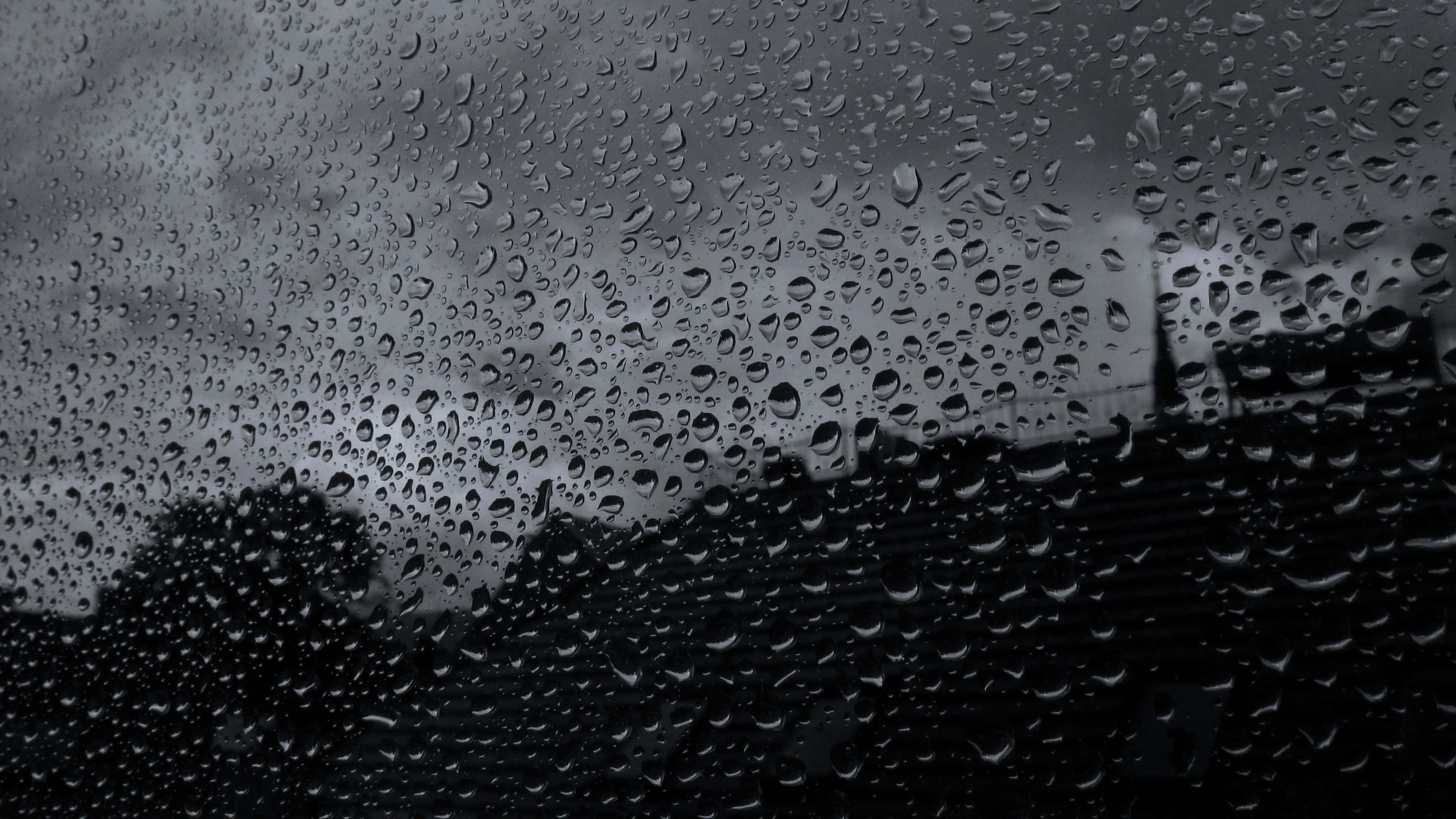 下雨 桌布 壁紙 — 雨滴依附在窗戶上，顯露出陰暗多雲的天空和遠處建築物的模糊輪廓。