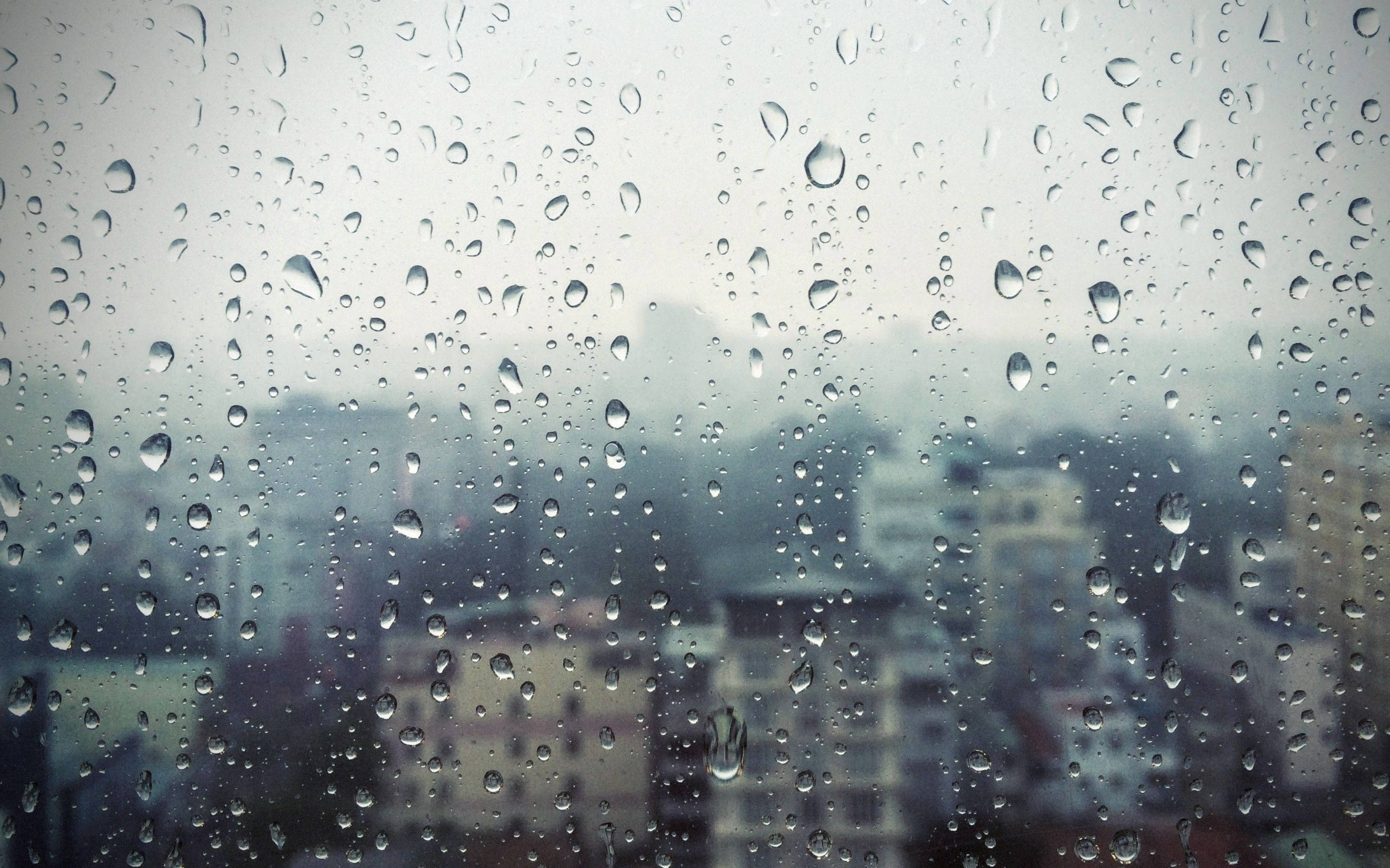 下雨 桌布 壁紙 — 雨滴依附在窗玻璃上，模糊了灰濛濛的城市天際線，呈現柔和的灰色和藍色。