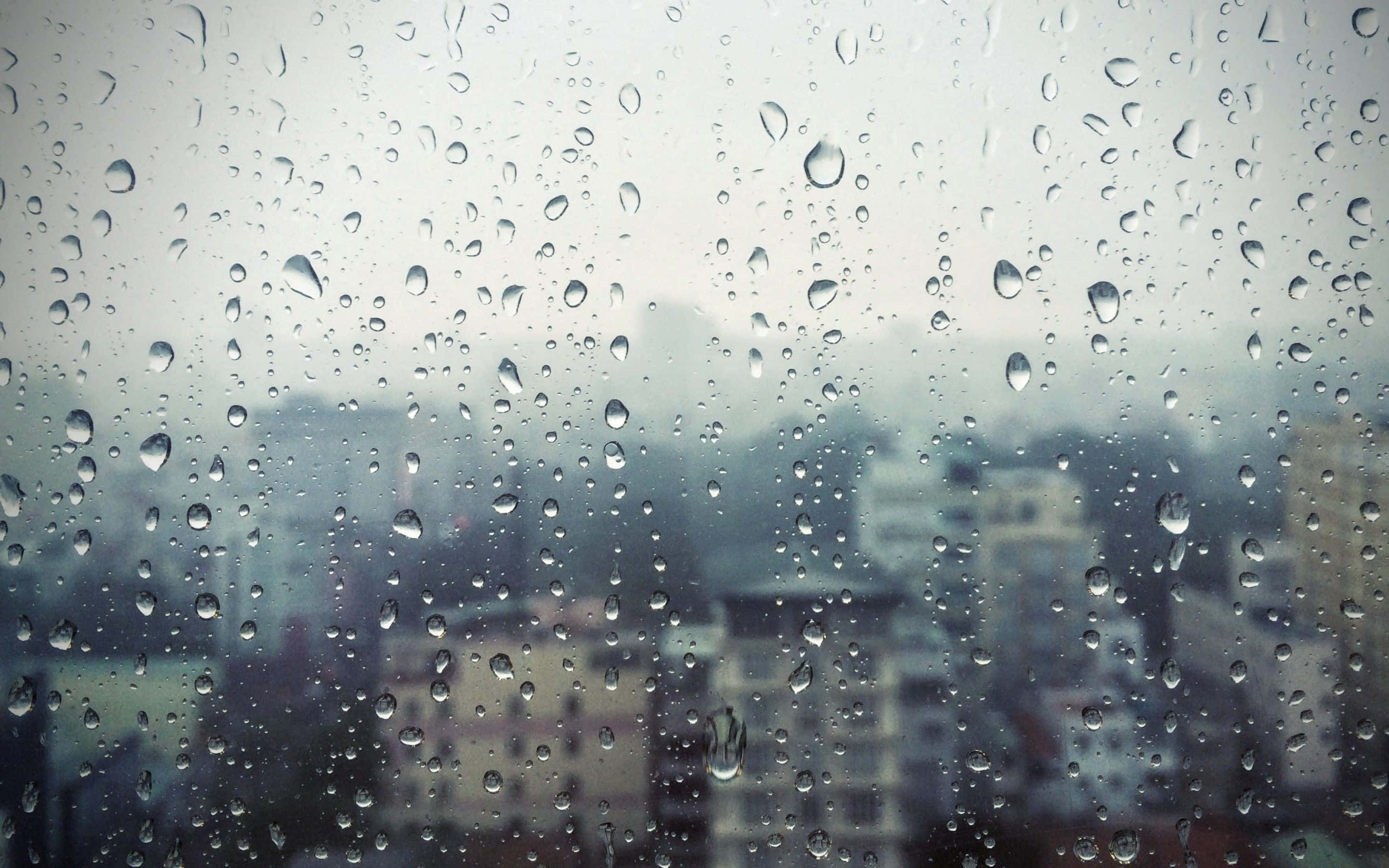Raining 桌布 壁紙 — 雨滴依附在窗玻璃上，模糊了陰天籠罩下的朦朧城市天際線。