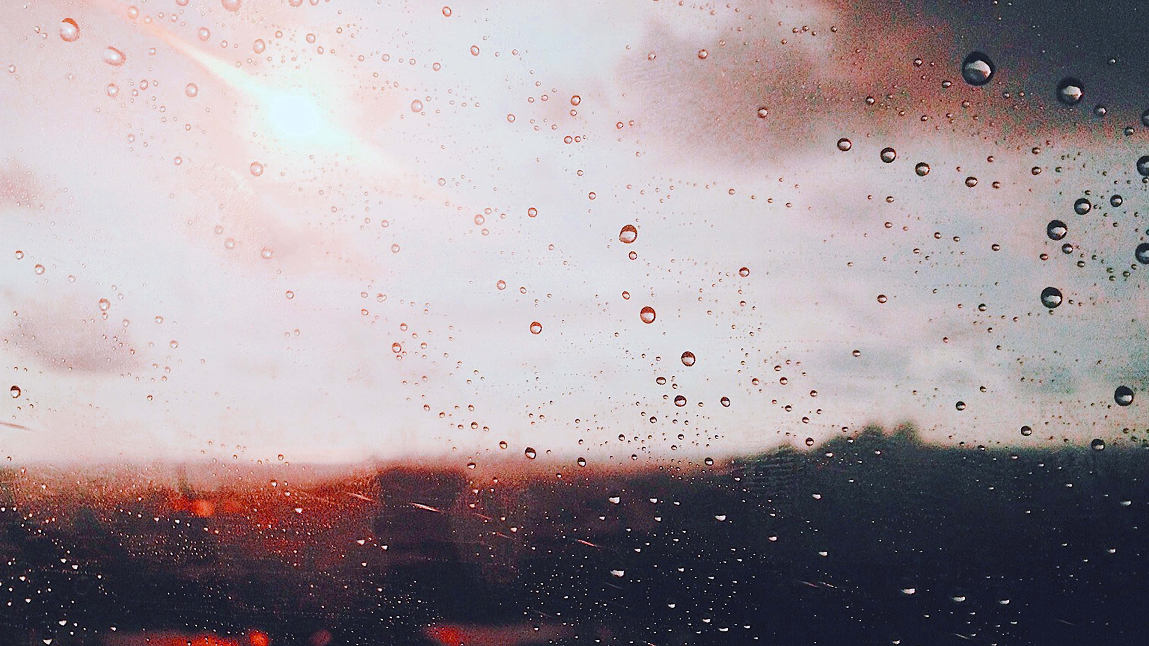 Rain 4K 桌布 壁紙 — 雨滴劃過窗戶，柔和的夕陽將模糊的城市景觀沐浴在溫暖的琥珀色和冷灰色調中。