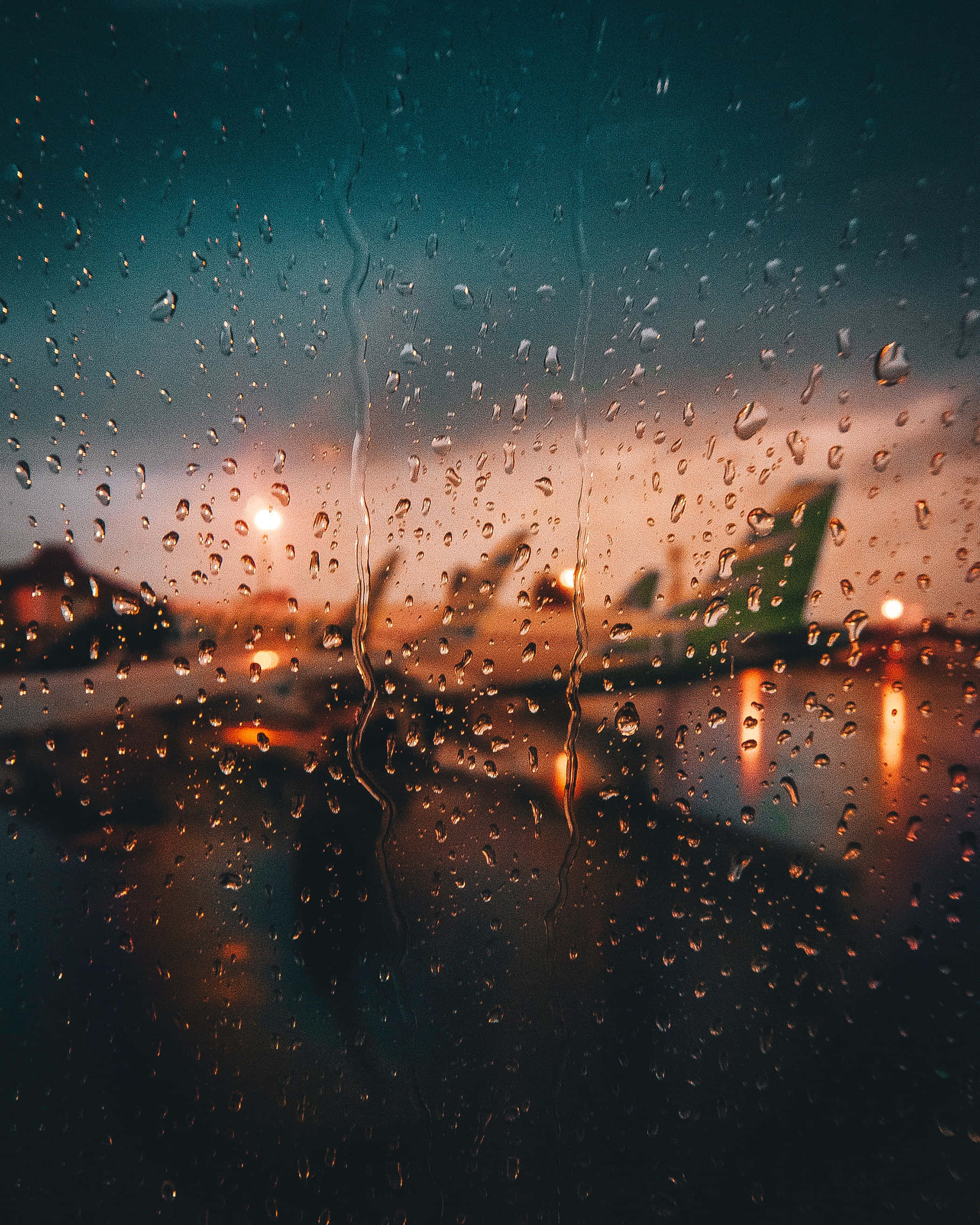 Raindrops 桌布 壁紙 — 雨滴劃過窗戶，顯露出暮色天空下溫暖燈光中模糊的機場飛機。