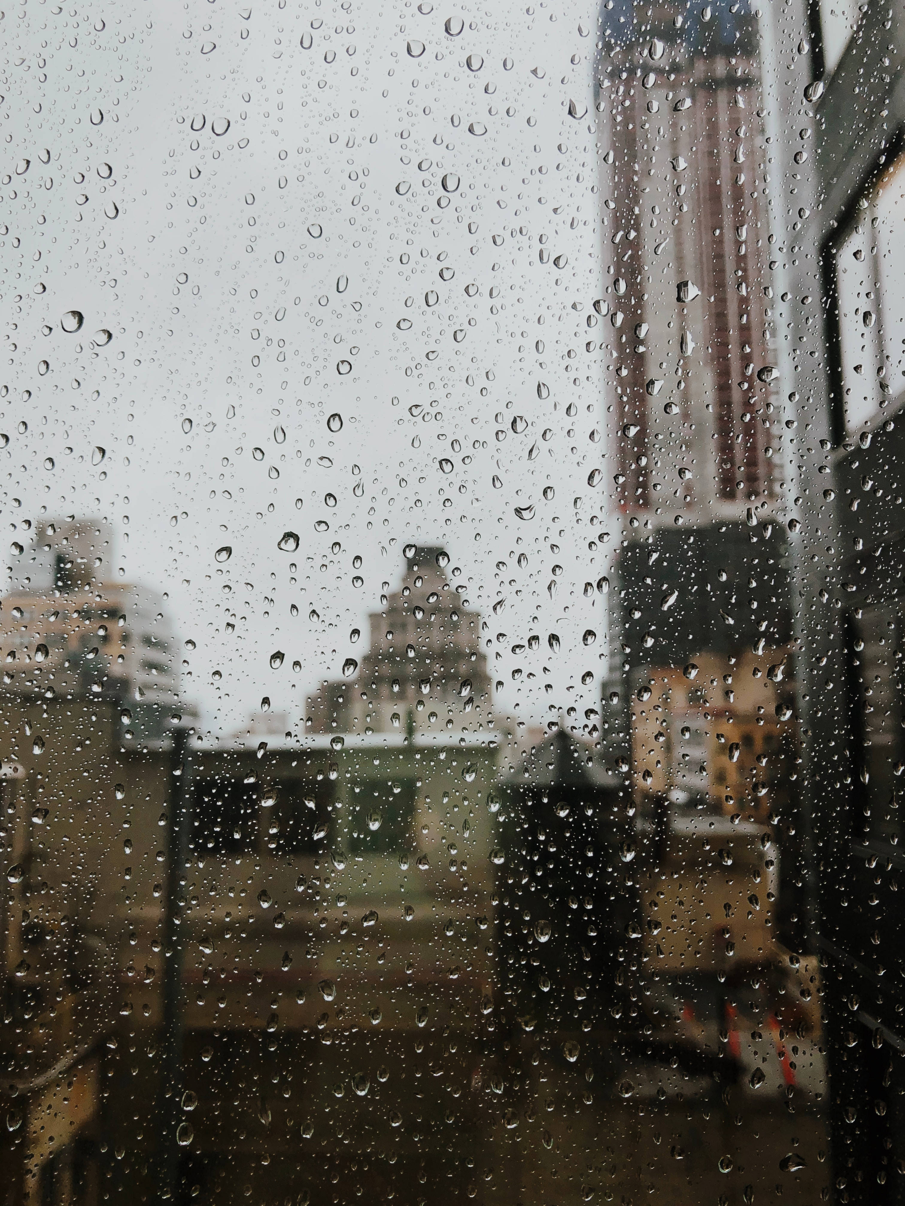 下雨 桌布 壁紙 — 雨滴劃過窗戶，顯露出陰天天空下模糊的城市景觀。
