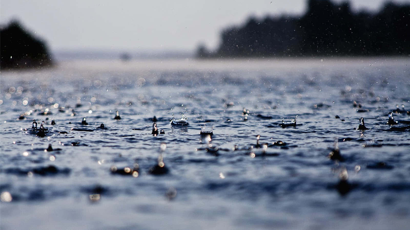 水 桌布 壁紙 — 雨滴在平靜的藍色水面上濺起漣漪，遠處的陸地在柔和日光下顯得模糊。