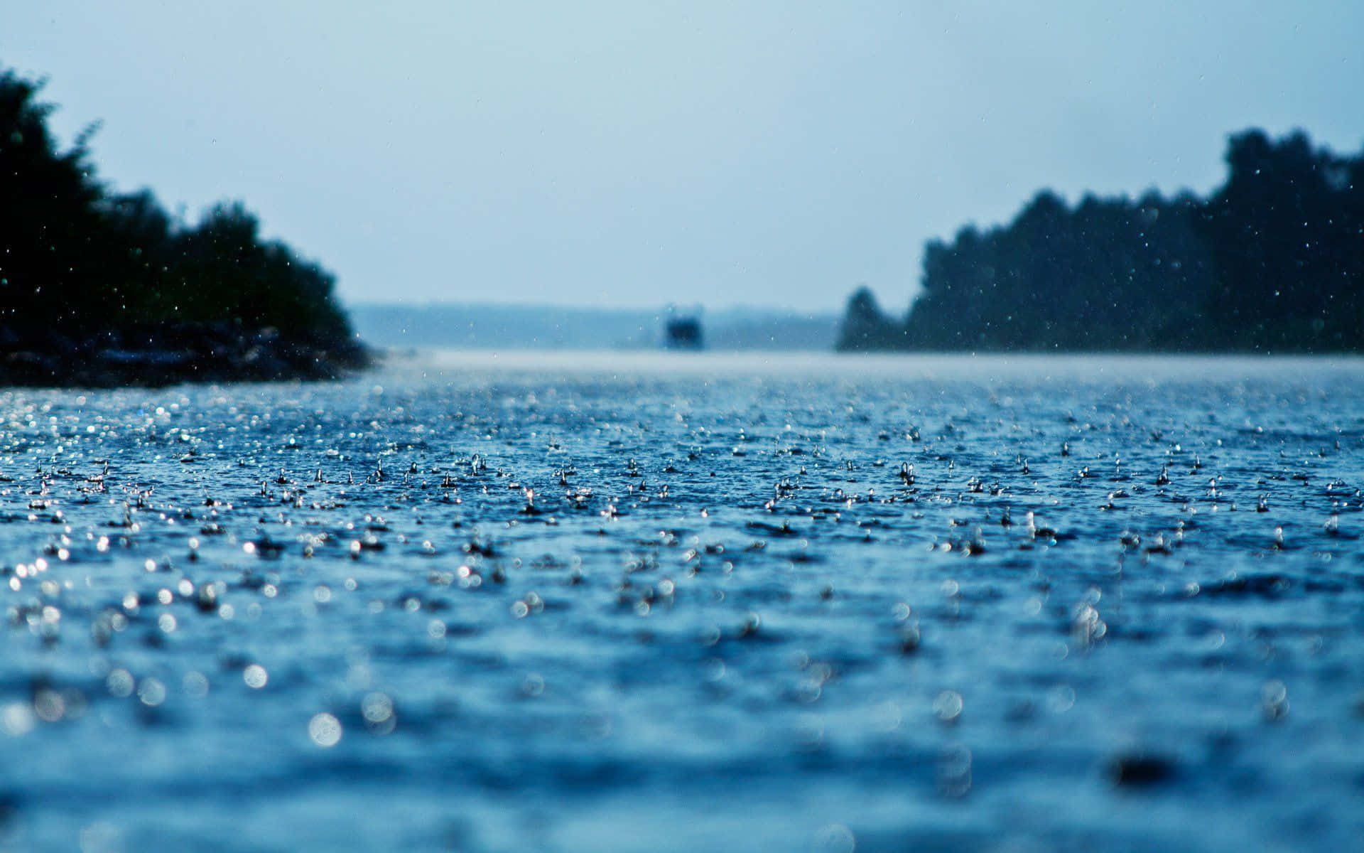 Free 桌布 壁紙 — 雨滴在廣闊的藍色湖面上泛起漣漪，天空朦朧，遠處有樹木成排的湖岸。