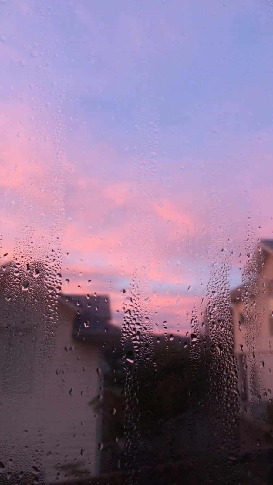 Aesthetic Rain 桌布 壁紙 — 雨滴在窗上劃過，柔和的粉紅與紫色夕陽在模糊的屋頂後方塗抹天空。