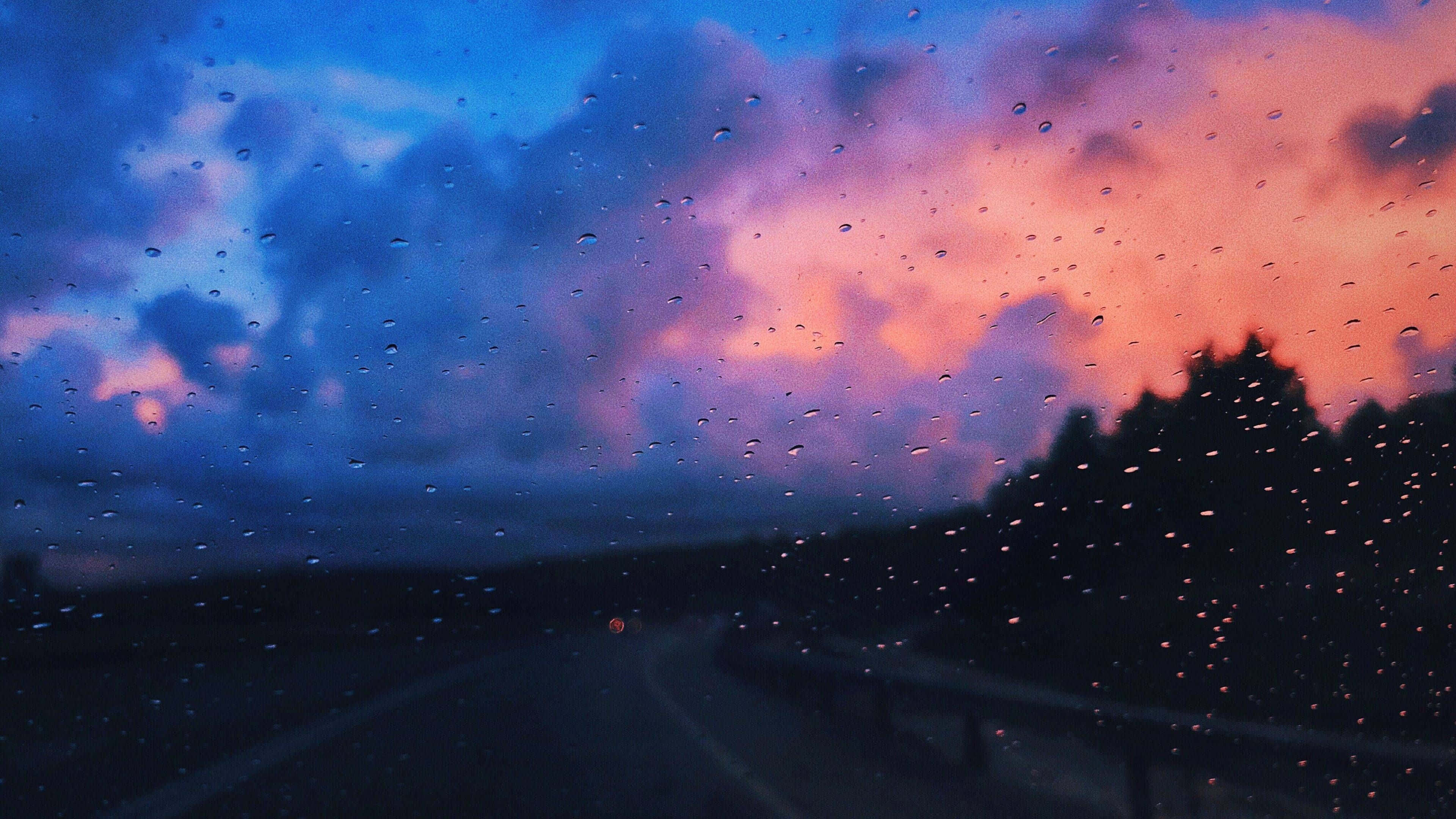 Nature Blur 桌布 壁紙 — 雨滴在車窗上劃出條紋，一條蜿蜒的道路在暮色天空下蜿蜒，天空被火橙色和深藍色的雲彩塗抹。