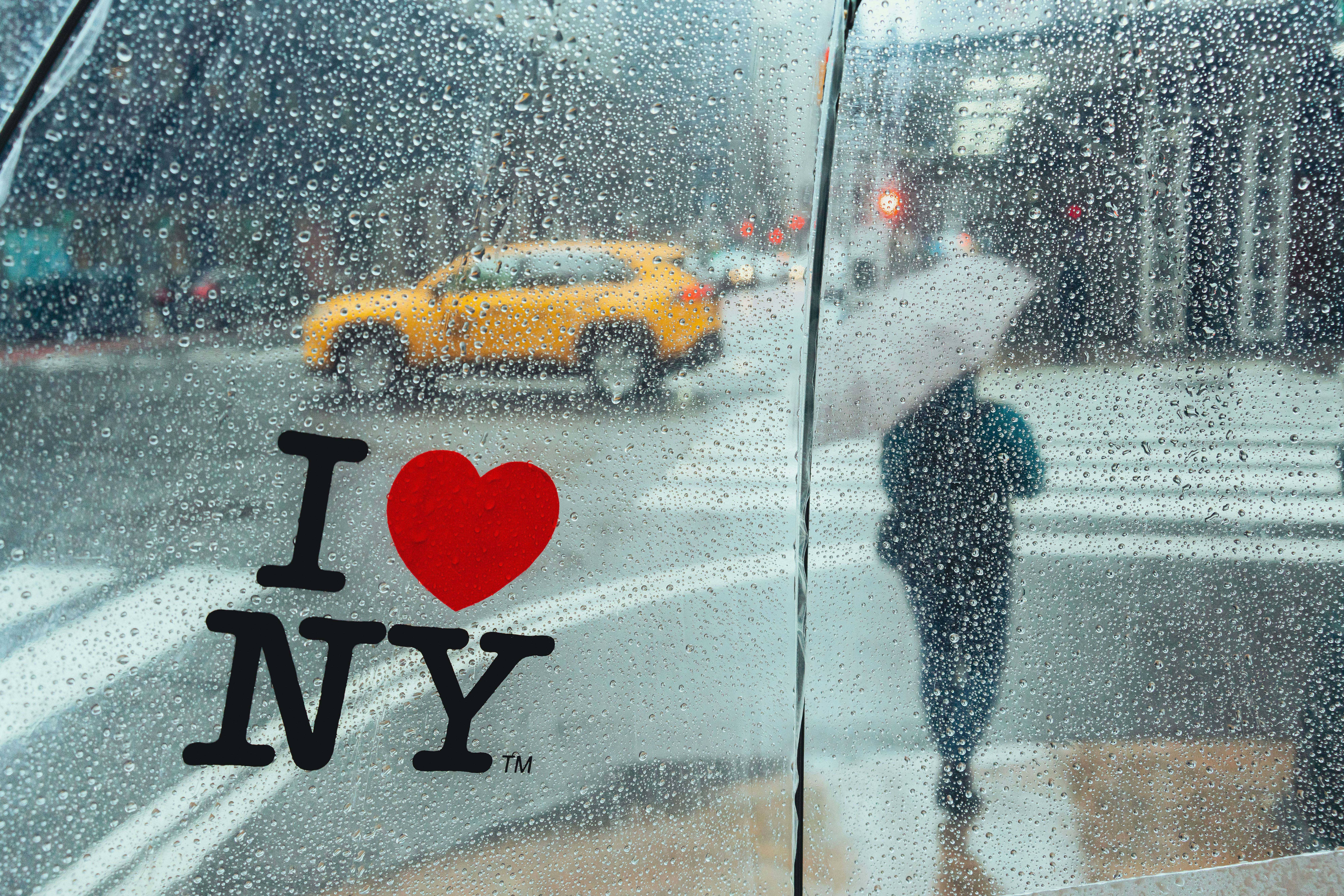 Nyc Aesthetic 桌布 壁紙 — 雨滴模糊了從車內看到的城市街景，車窗上貼有 I heart NY 貼紙，一輛黃色計程車駛過。