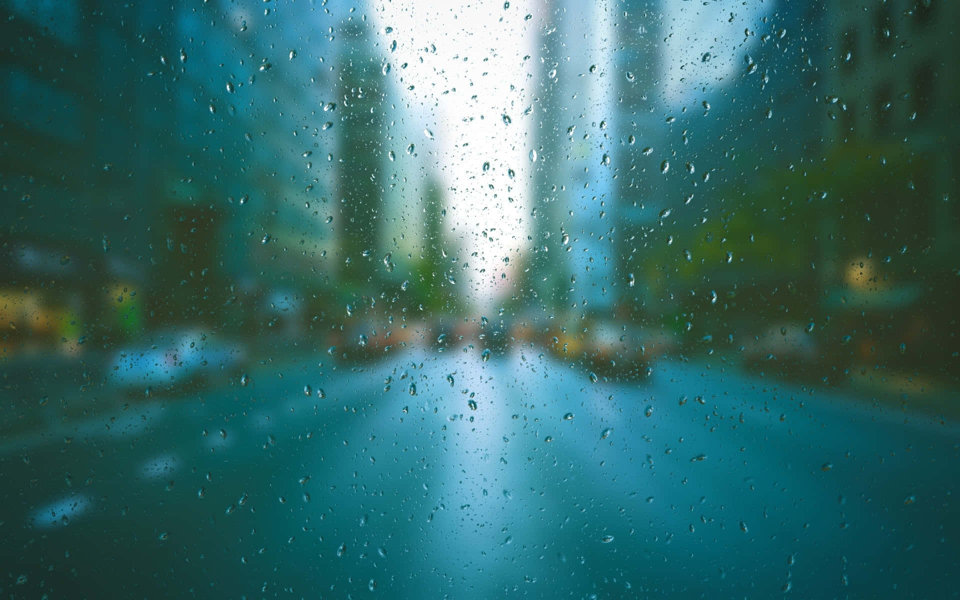 images 桌布 壁紙 — 雨滴模糊了透過窗戶看到的城市街道，遠處高樓大廈柔和地發光。