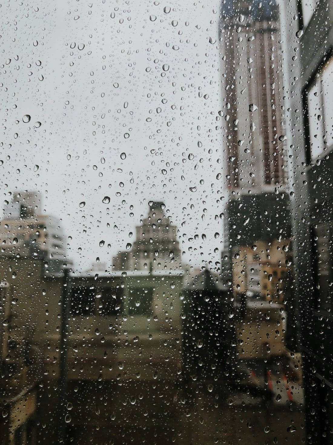 Rainy Day 桌布 壁紙 — 雨滴沿窗戶流下，顯露出陰天天空下模糊的城市景觀。
