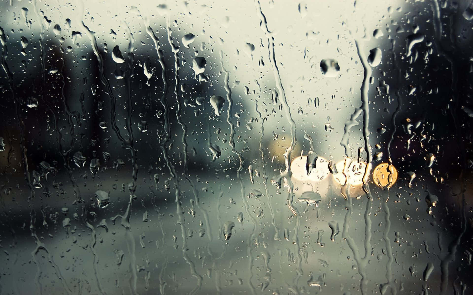 Raining 桌布 壁紙 — 雨滴沿著玻璃窗流下，將城市燈光模糊成柔和發光的光球，映襯在灰色背景上。