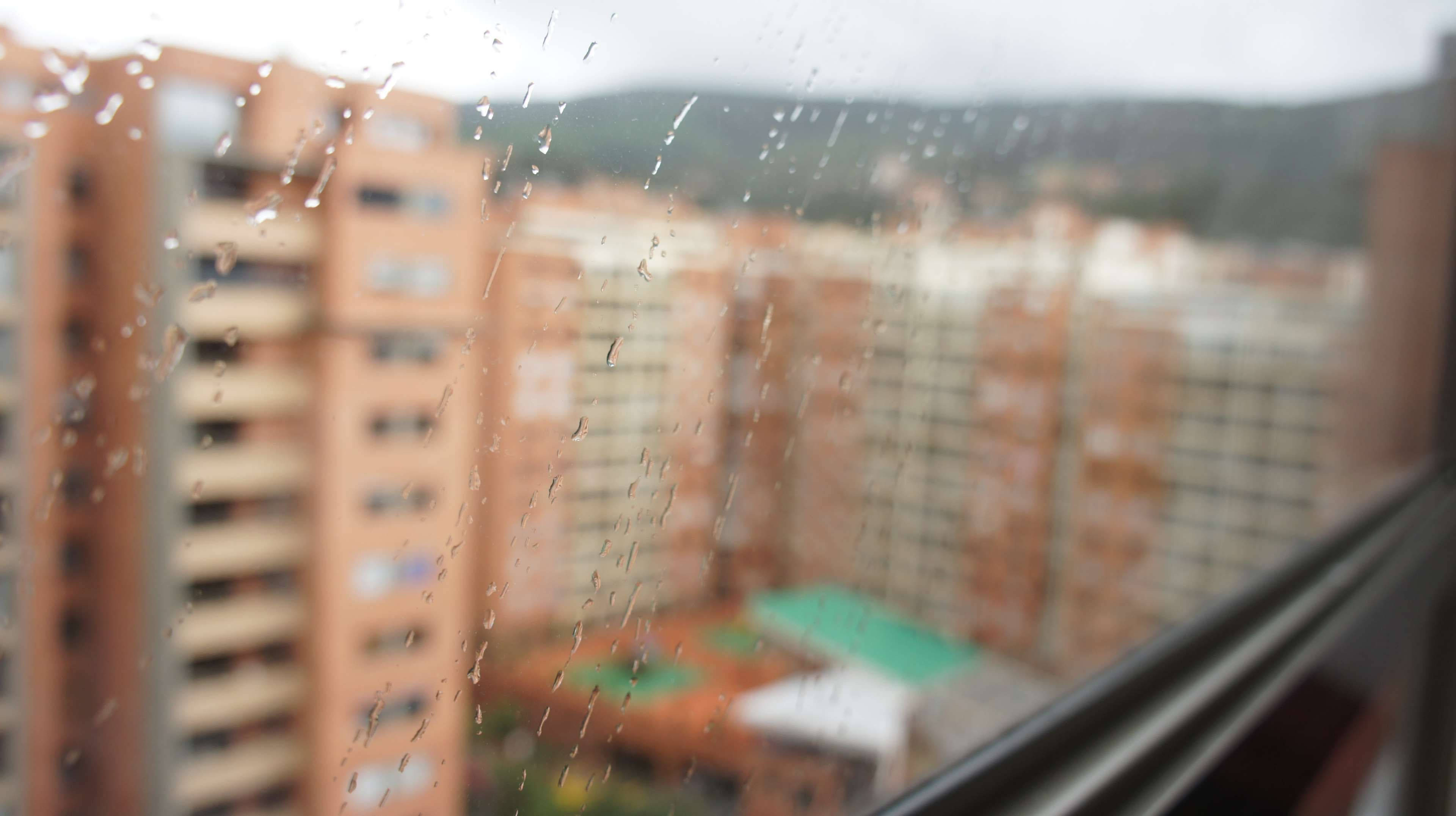 Bogota 桌布 壁紙 — 雨滴沿著窗玻璃流下，模糊了陰天天空下橙色公寓樓和遠處綠色山丘的景色。