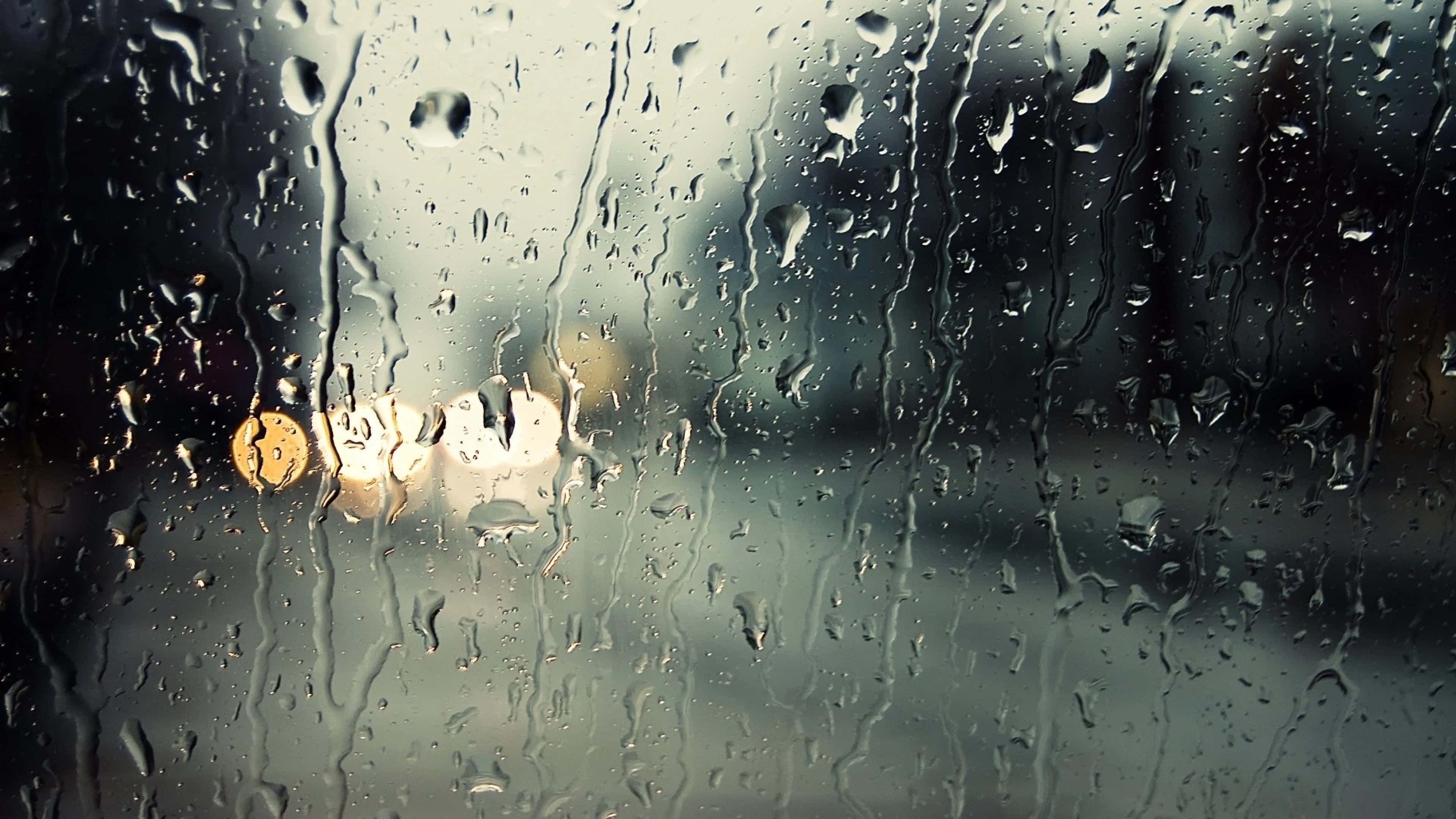 下雨 桌布 壁紙 — 雨滴沿著車窗流下，將溫暖的街燈模糊成發光球體，映襯著灰色的城市背景。