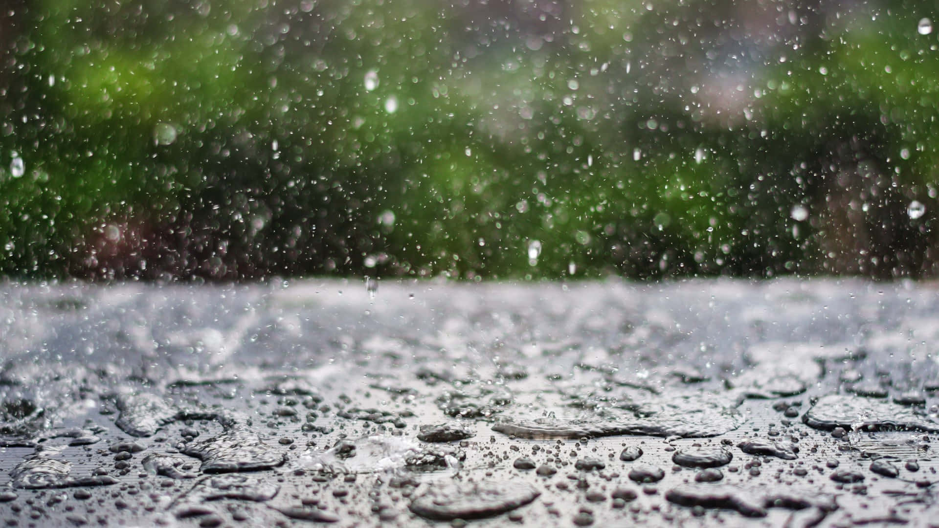 下雨 桌布 壁紙 — 雨滴濺起並在潮濕的表面上形成水珠，背景是模糊的綠色樹葉。