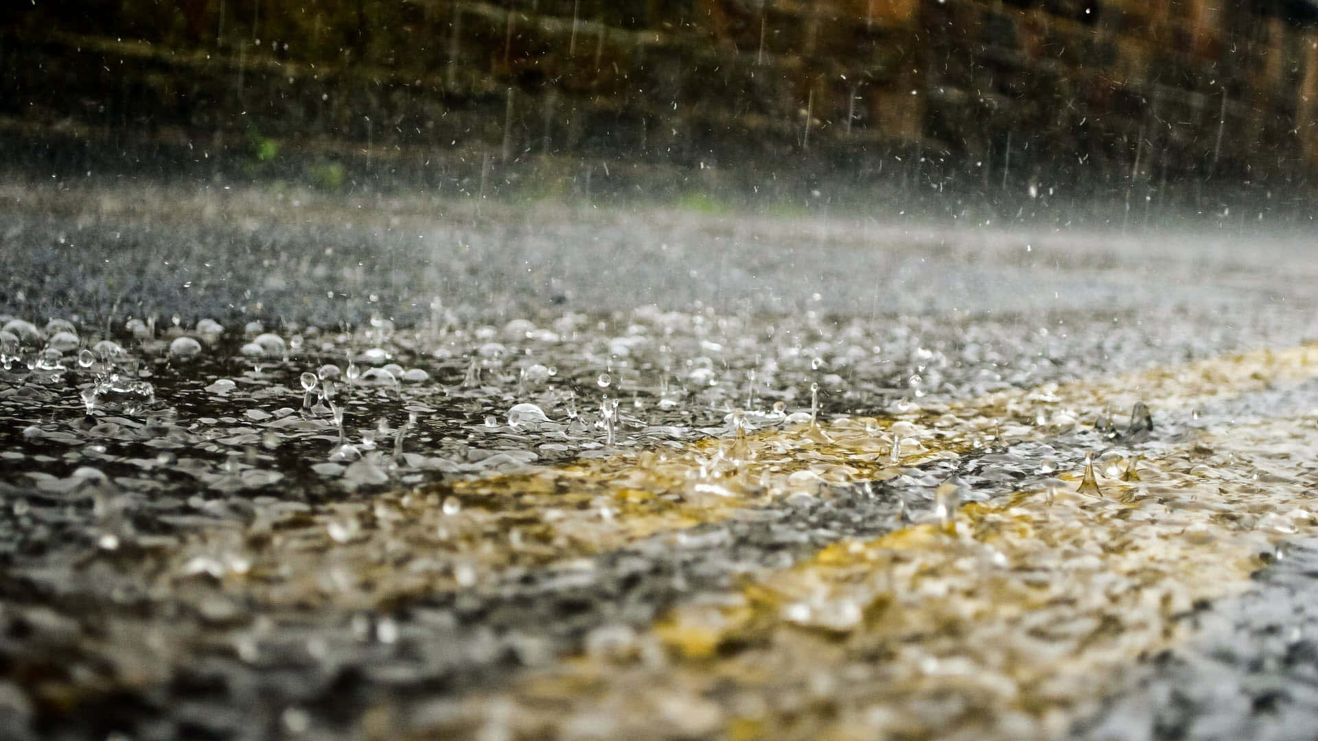 下雨 桌布 壁紙 — 雨滴濺起並在濕漉漉的瀝青路上泛起漣漪，路上有一條褪色的黃線。