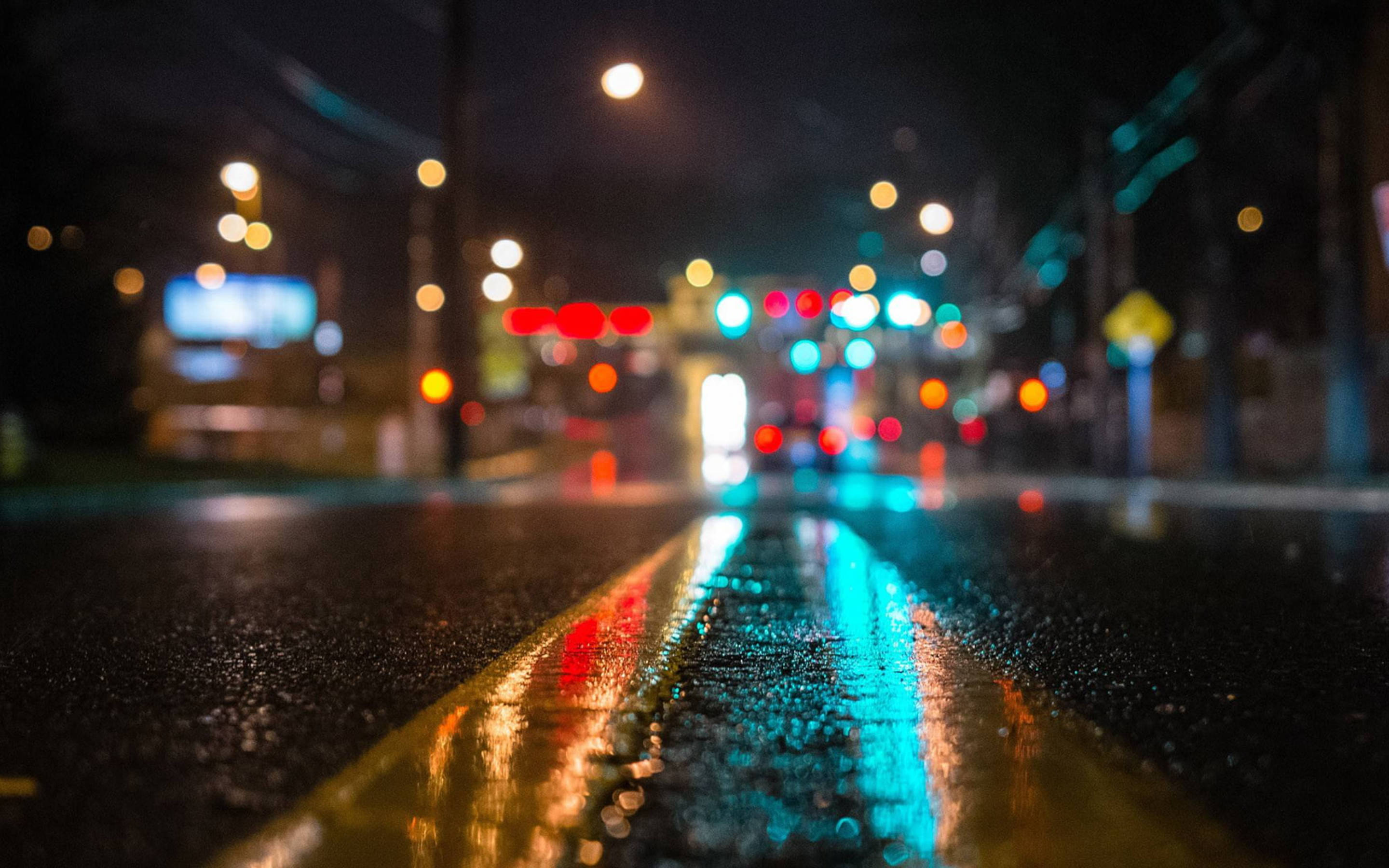 Modern 桌布 壁紙 — 雨濕的夜晚城市街道，濕漉漉的柏油路和黃色車道標記上反射著發光的散景燈光。