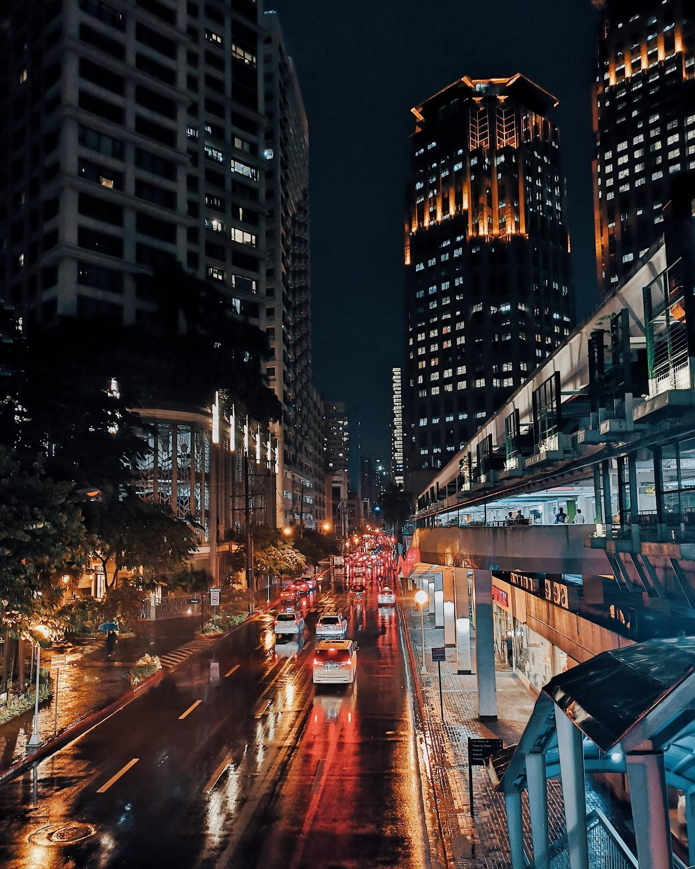 Manila 桌布 壁紙 — 雨濕的夜晚城市街道，發光的汽車尾燈在濕漉漉的路面上反射，兩旁是高聳的照明摩天大樓。