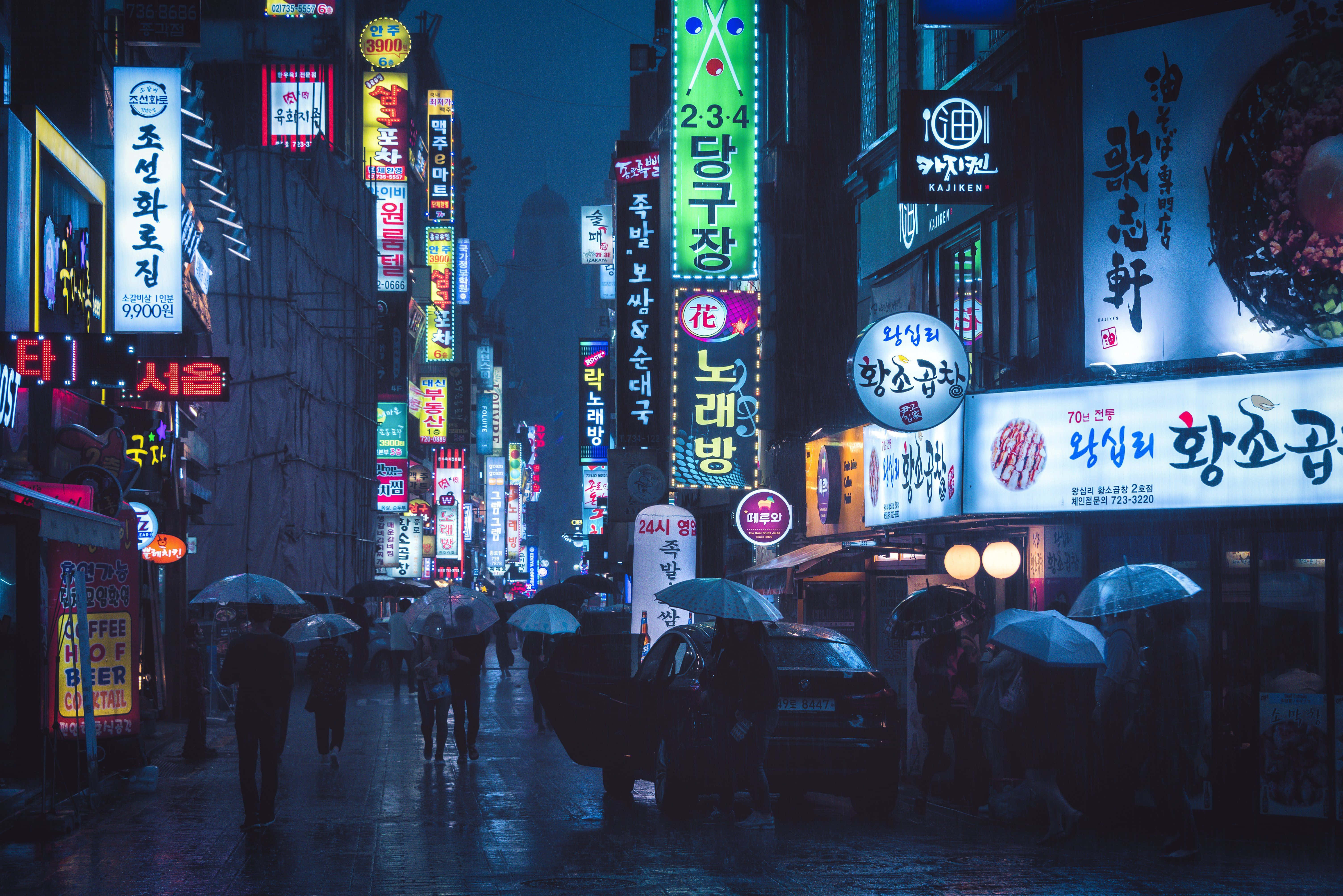 桌布 壁紙 — 雨濕的夜晚城市街道，閃爍著鮮豔的霓虹招牌和撐傘的行人。