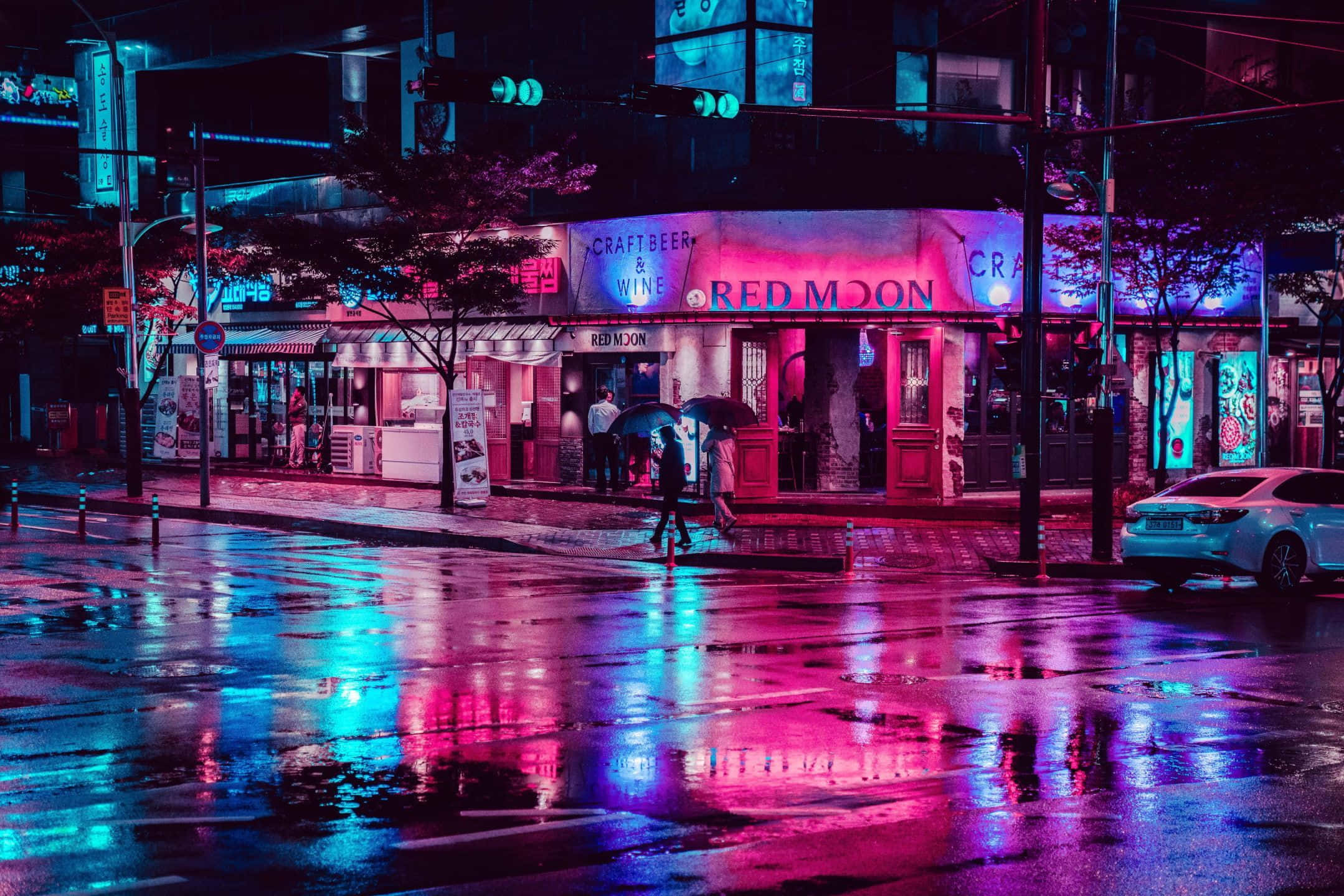 霓虹城市動態圖 桌布 壁紙 — 雨濕的夜晚城市街道反射著商店櫥窗的霓虹粉紅和藍色燈光，行人撐著傘穿過濕漉漉的人行道。