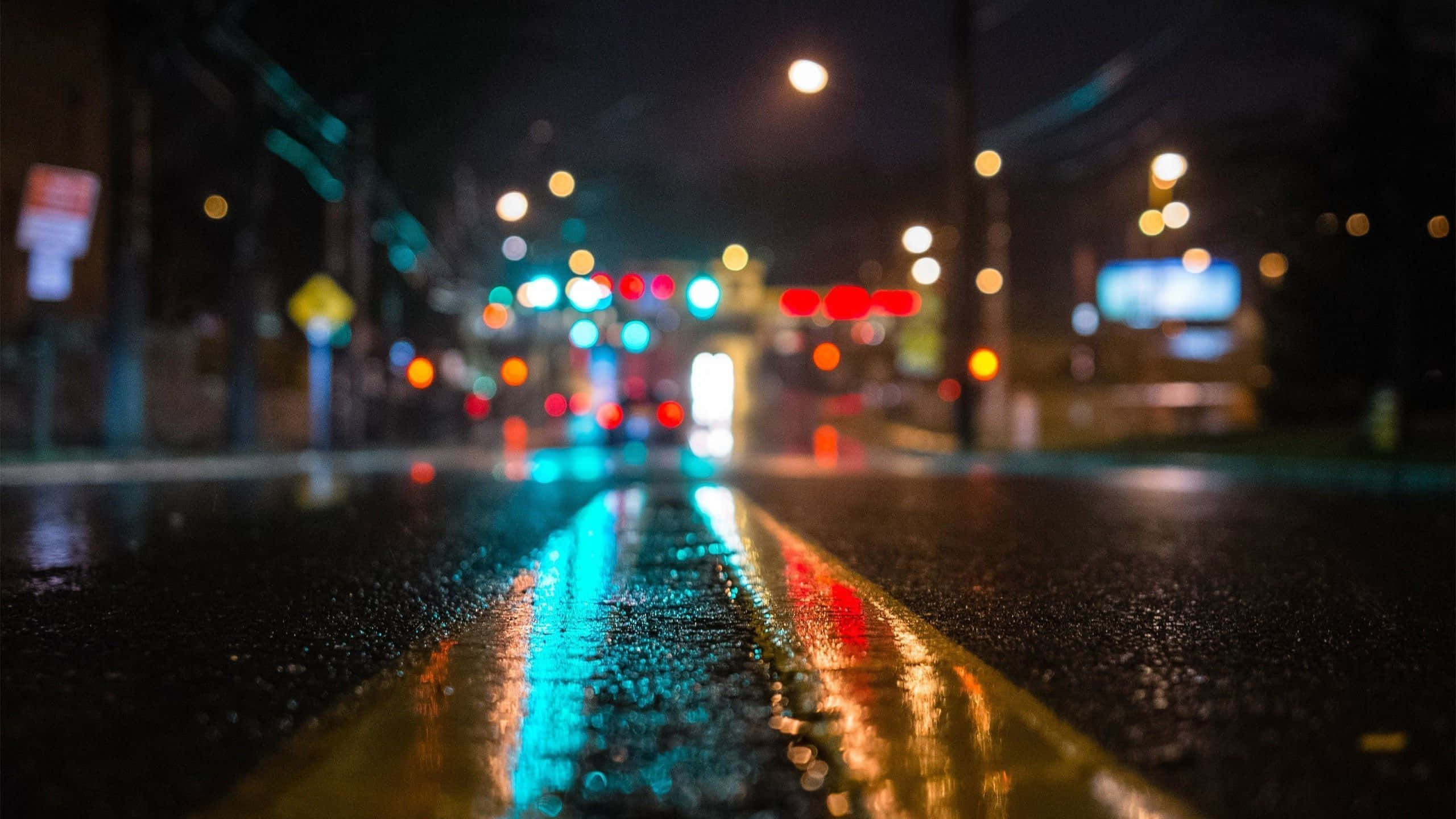 2560x1440 動態圖片 桌布 壁紙 — 雨濕的夜間城市街道，發光的交通燈反射在濕瀝青上，背景是模糊的城市燈光。