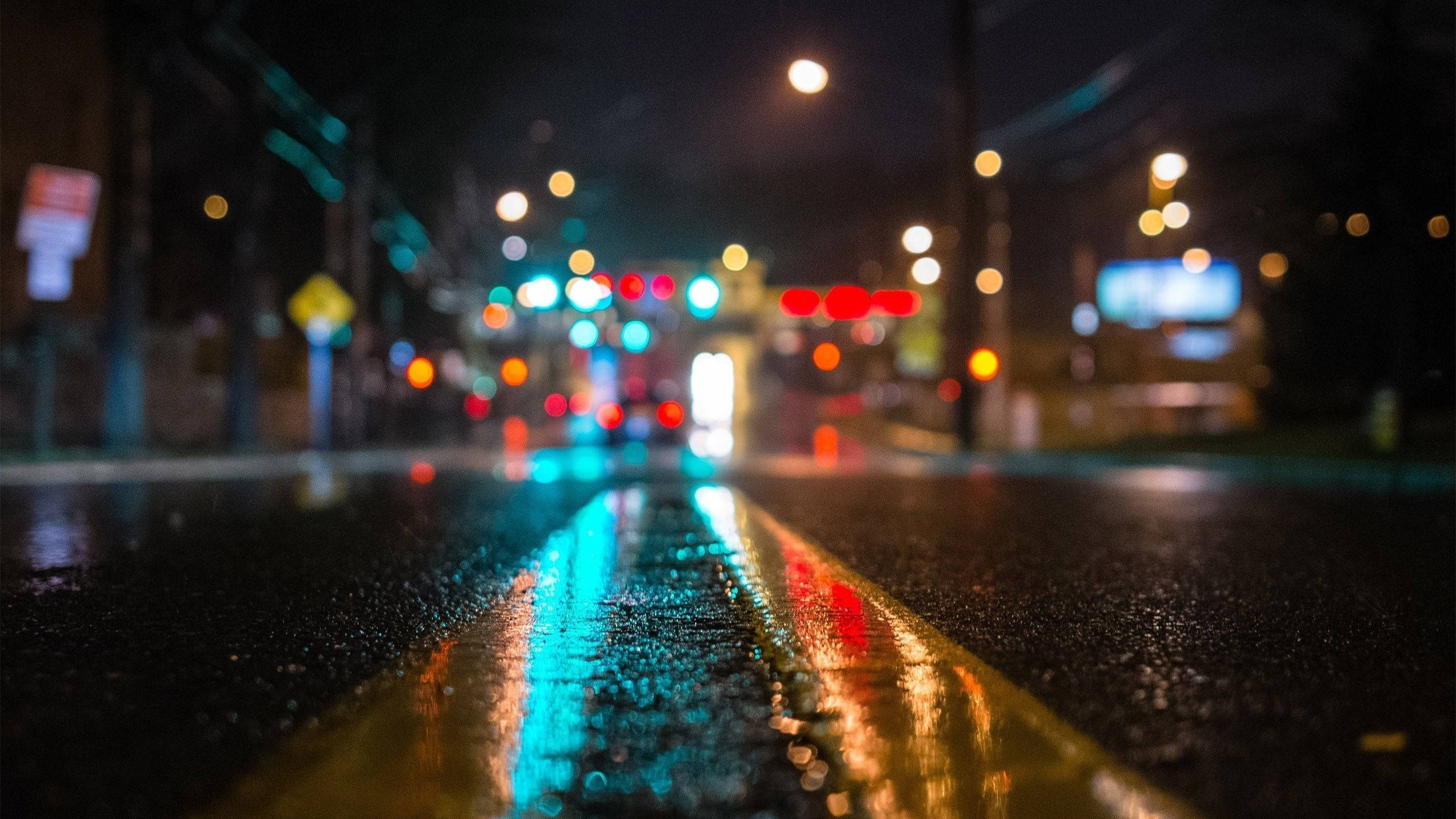 Street 桌布 壁紙 — 雨濕的夜間城市街道，發光的散景燈光反射在潮濕的瀝青和黃色車道線上。