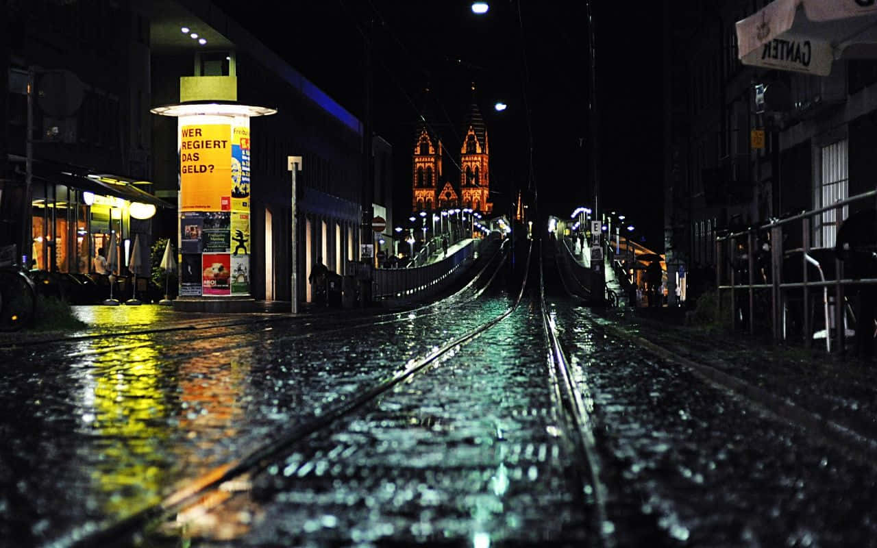 Rainy 桌布 壁紙 — 雨濕的夜間城市街道，發光的電車軌道通向燈火通明的教堂尖塔。