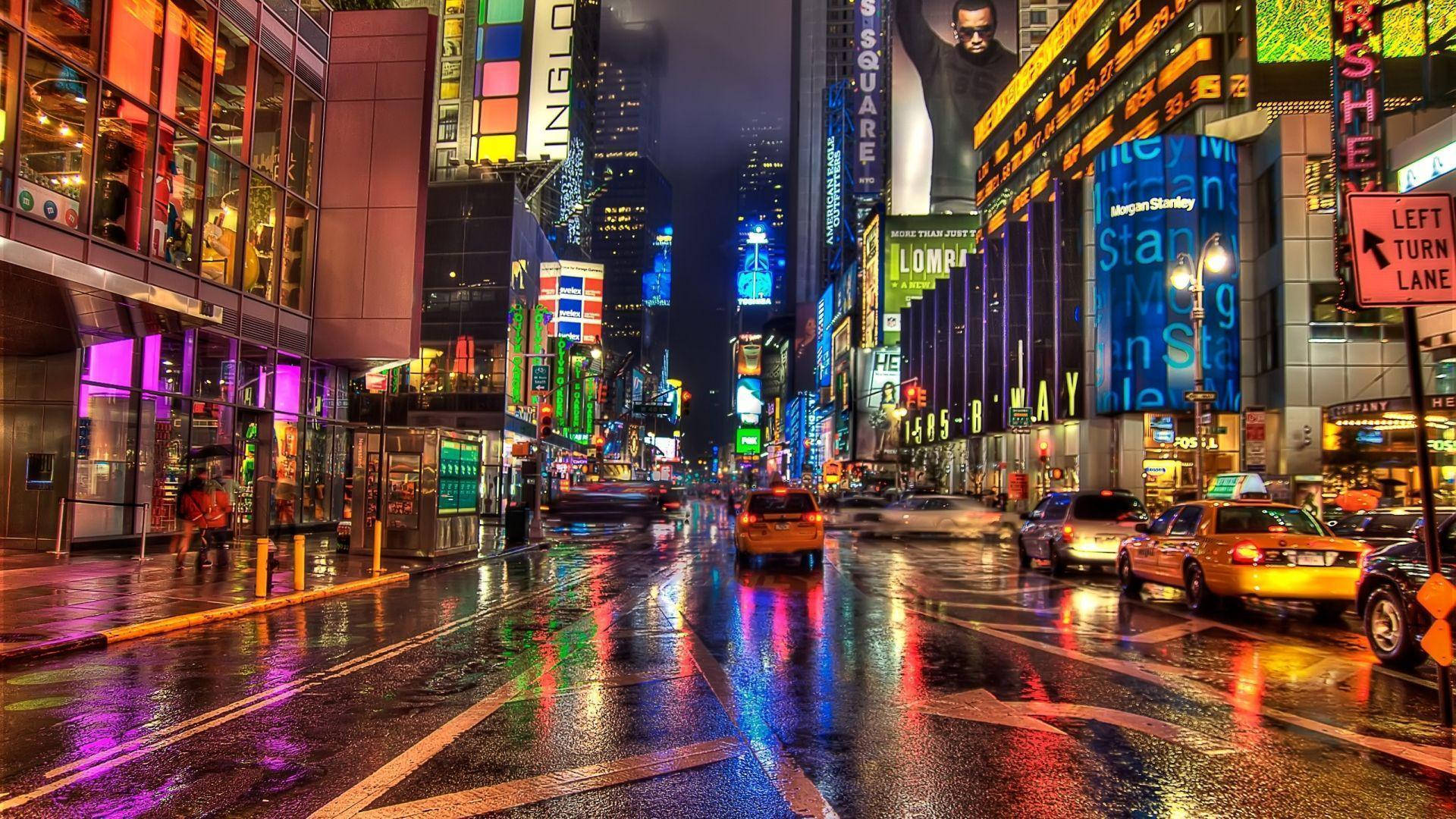 Broadway 桌布 壁紙 — 雨濕的時代廣場夜晚在高聳的數位廣告牌下，濕漉漉的路面上閃爍著霓虹燈的倒影。