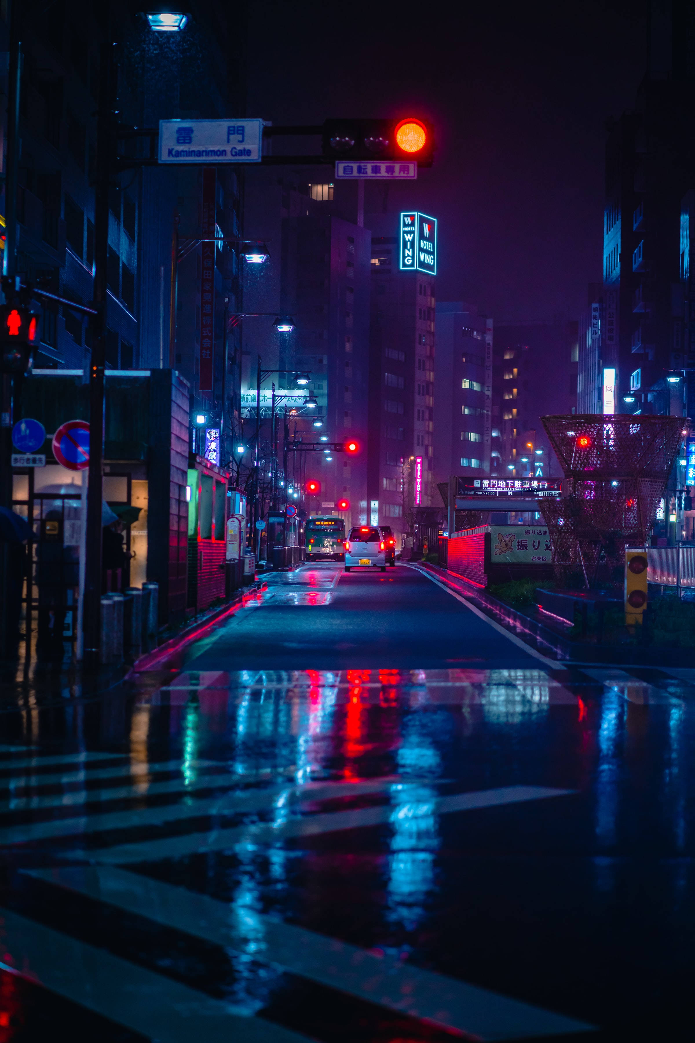 東京 桌布 壁紙 — 雨濕的東京夜晚街道，在黑暗的天空下反射霓虹燈牌和紅色交通燈。