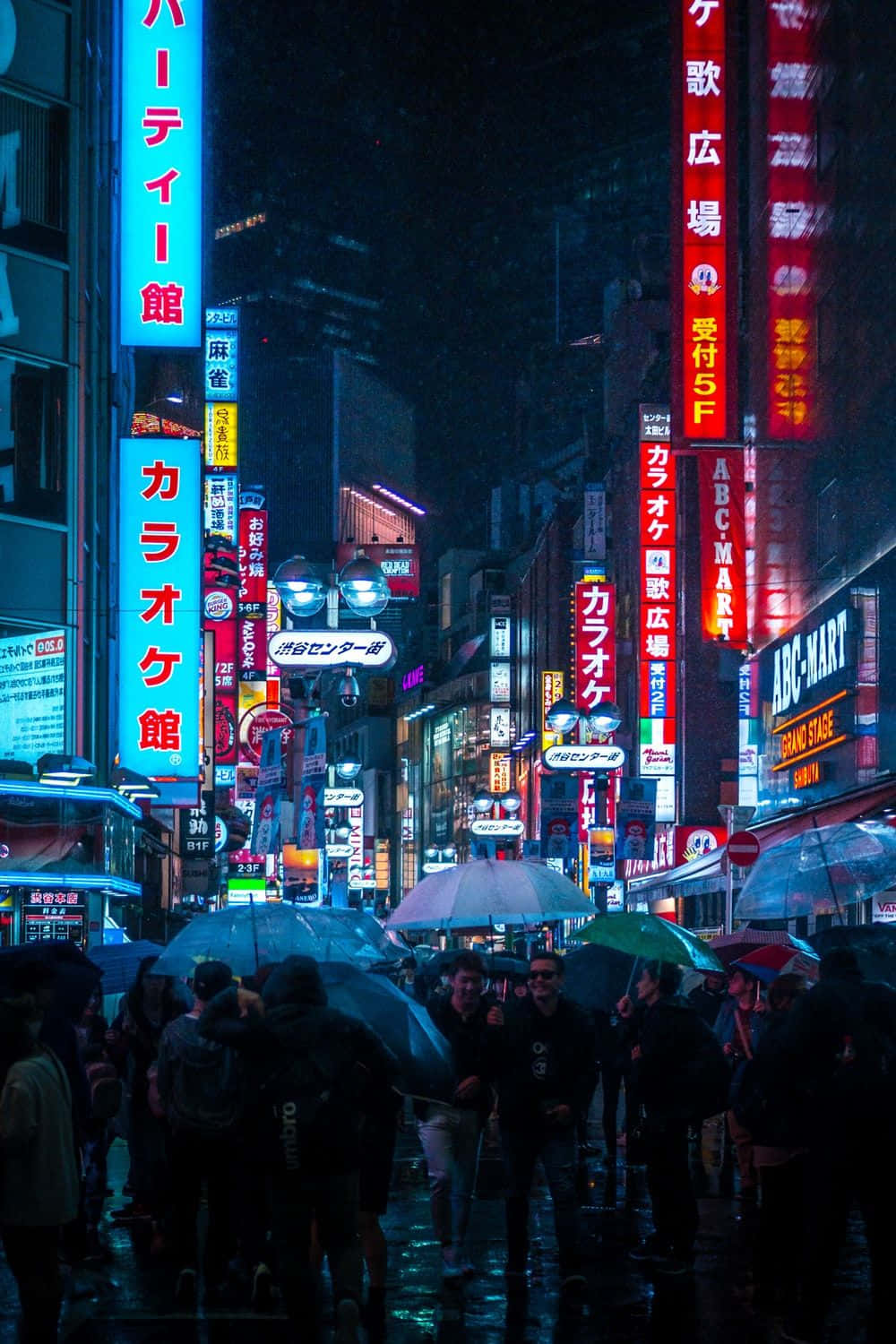 桌布 壁紙 — 雨濕的東京夜晚街道，霓虹招牌閃爍，人們在濕漉漉的行人路上撐傘。