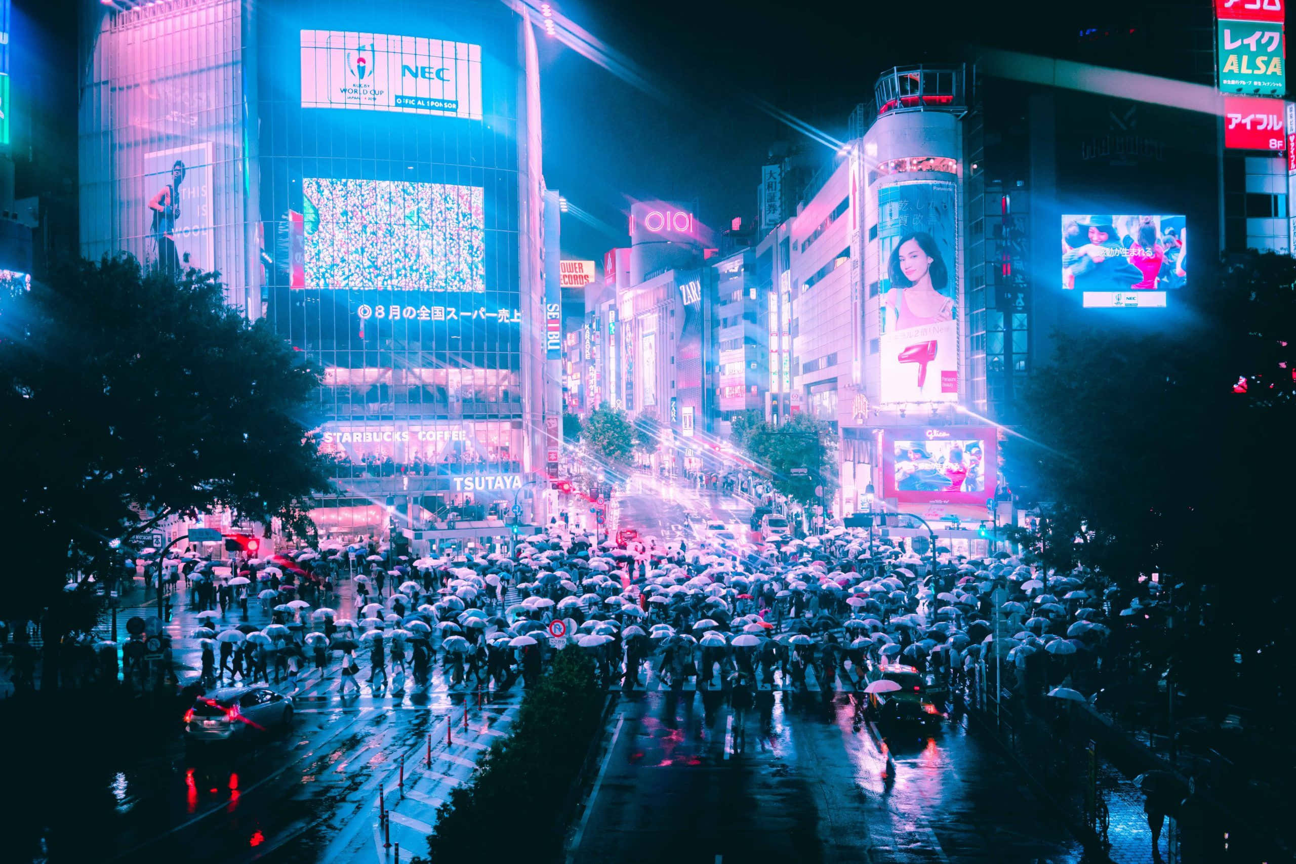 日本 桌布 壁紙 — 雨濕的東京夜晚街道，霓虹燈閃爍，人群在高聳的數位廣告牌下撐著傘。
