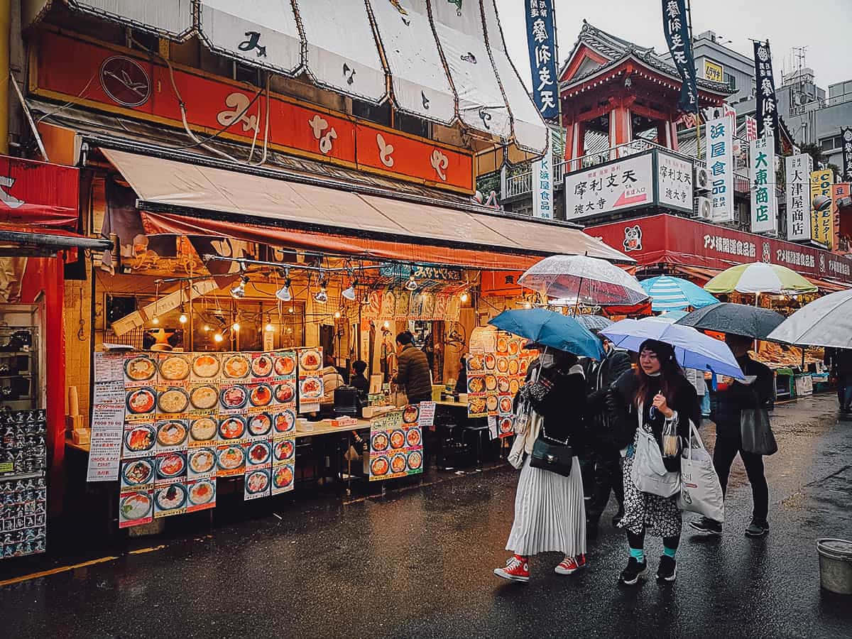 Street 桌布 壁紙 — 雨濕的東京街道，行人撐傘經過充滿活力的美食攤位和傳統紅色招牌。