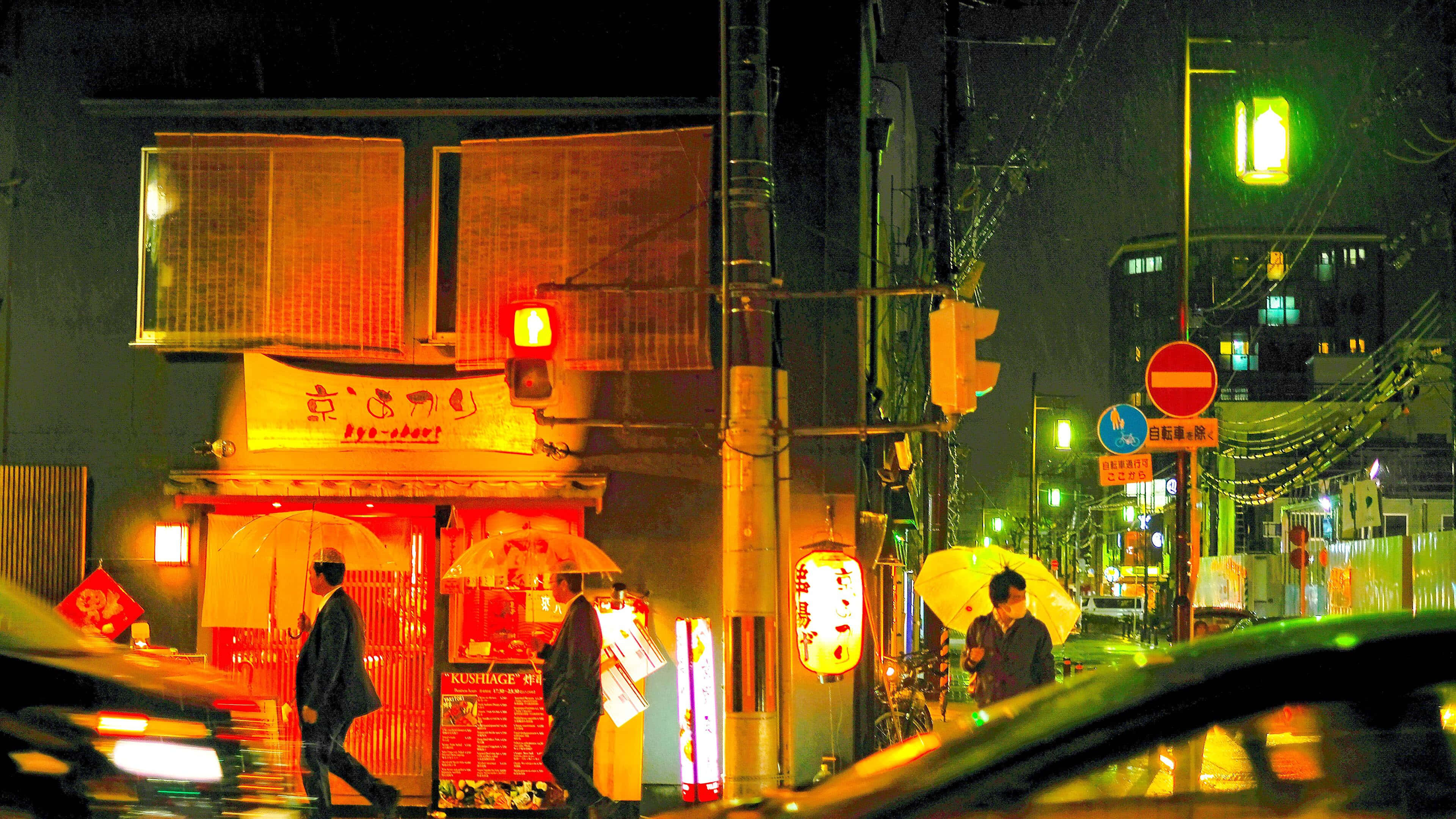 日本 桌布 壁紙 — 雨濕的東京街道在夜晚閃爍著霓虹燈和溫暖的燈籠，行人撐著傘在潮濕的城市小巷中穿行。