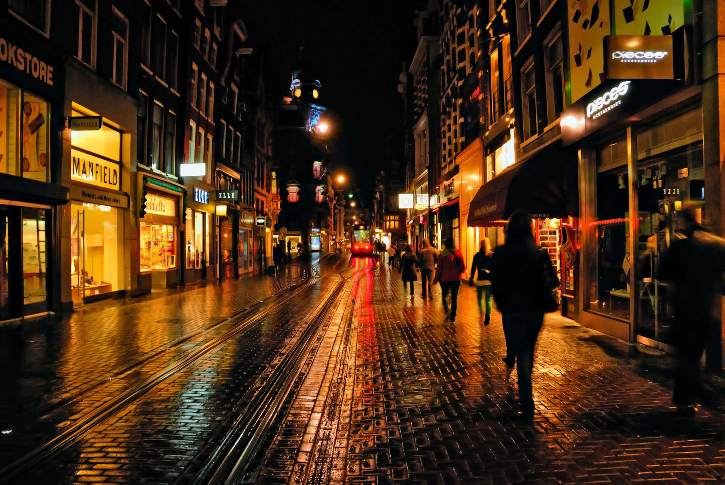 Night Street 桌布 壁紙 — 雨濕的鵝卵石街道夜晚，發光的商店櫥窗和模糊的行人在溫暖的路燈下行走。