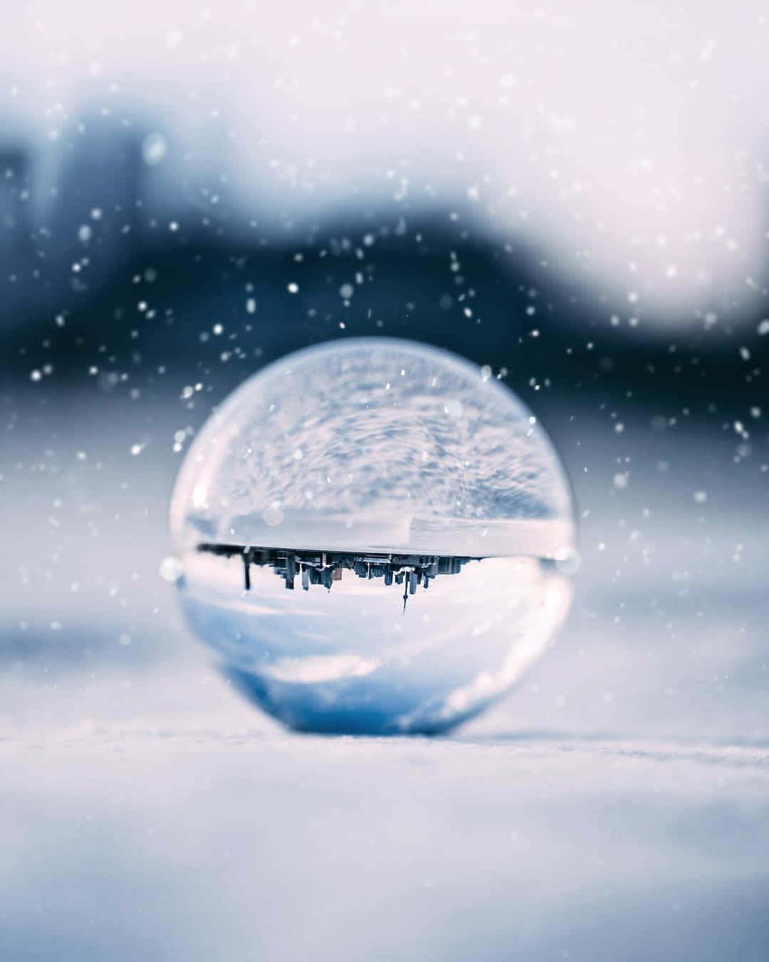 Crystal Ball 桌布 壁紙 — 雪地上清澈的水滴反射出在飄落的雪花下模糊的城市天際線，呈現冷藍色調。