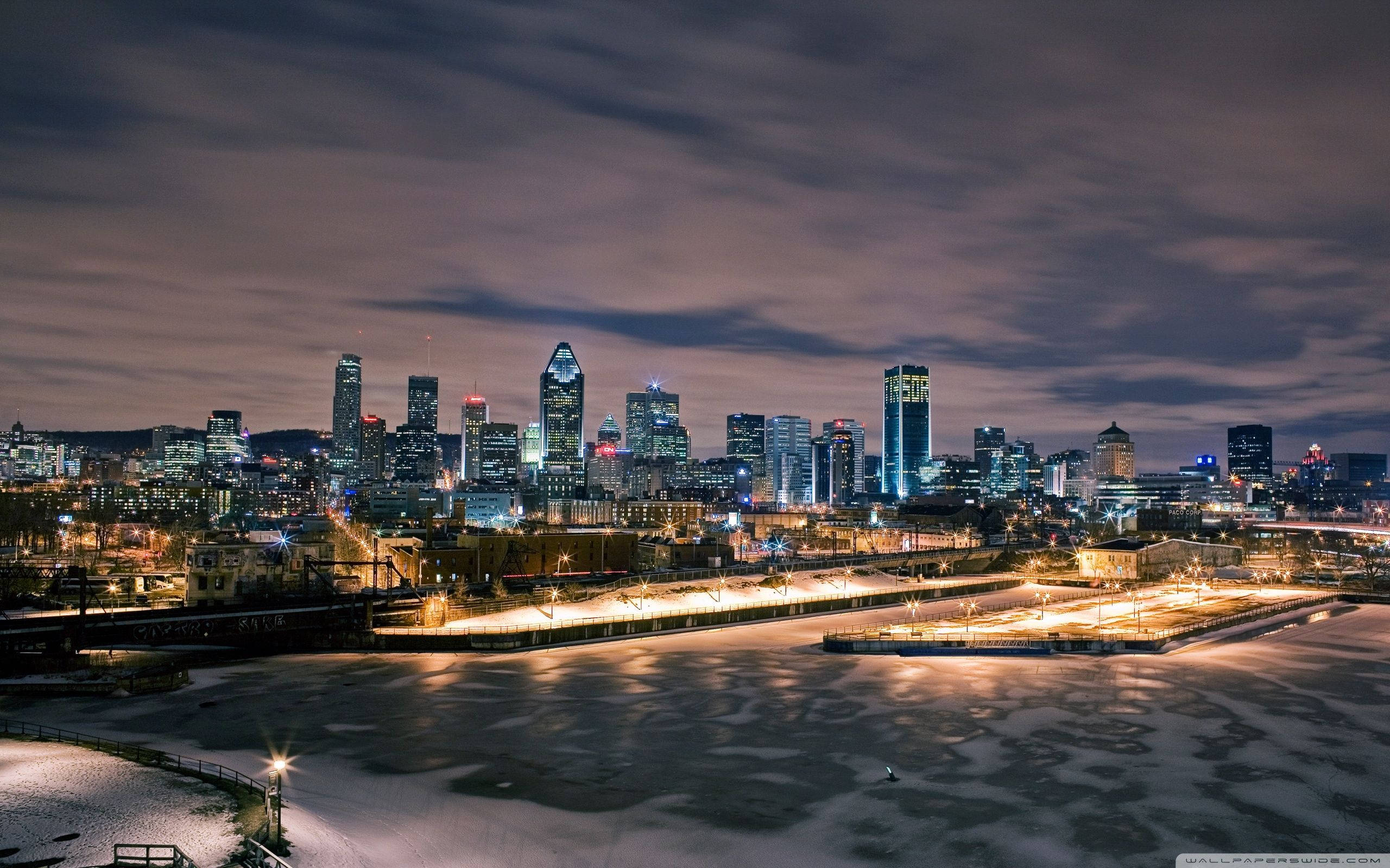 加拿大 桌布 壁紙 — 雪景城市夜景，在陰雲密布的暮色天空下，發光的摩天大樓倒映在結冰的河面上。