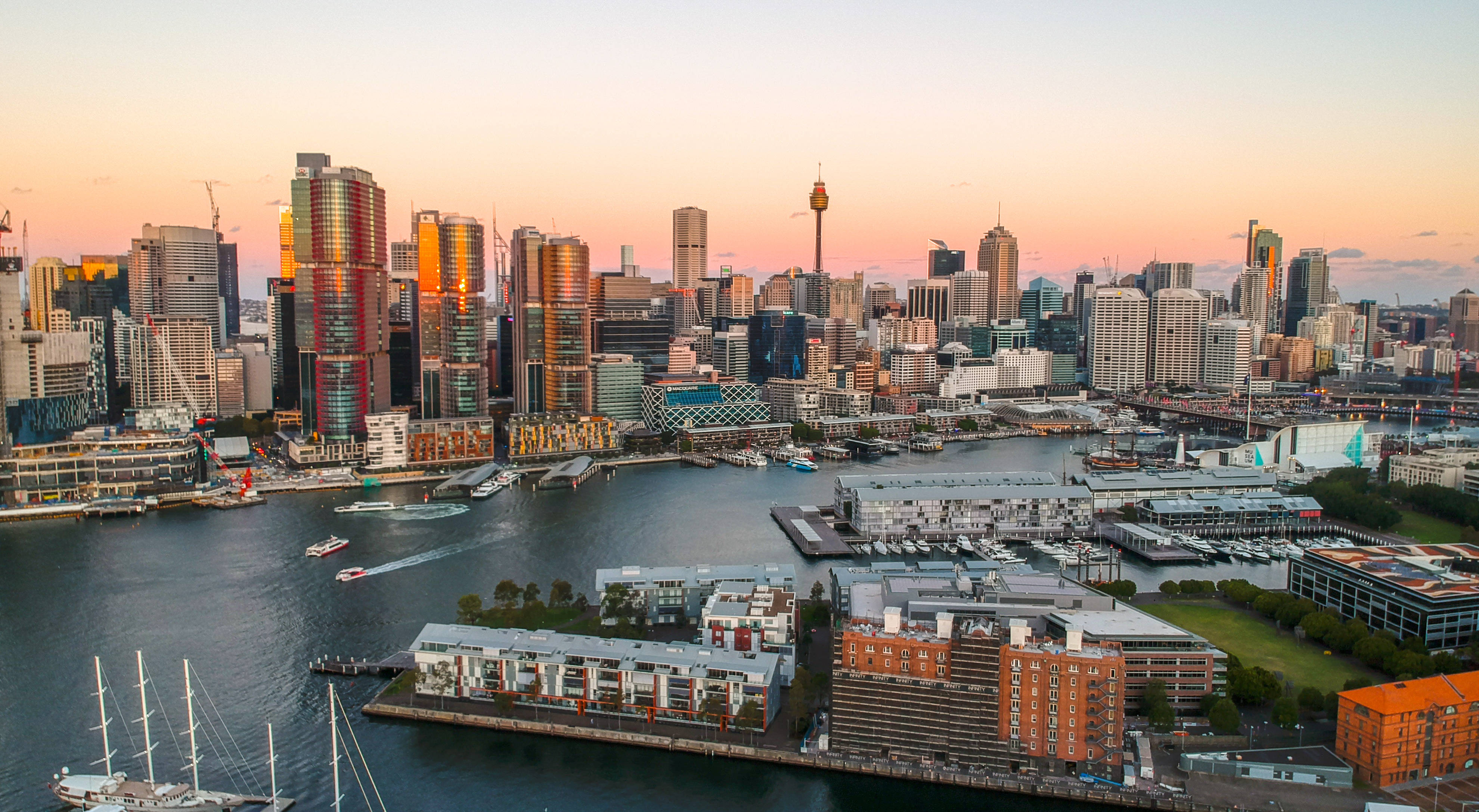 Sydney 桌布 壁紙 — 雪梨天際線的航拍圖，夕陽下金色摩天大樓倒映在港灣水面，下方停泊著帆船。