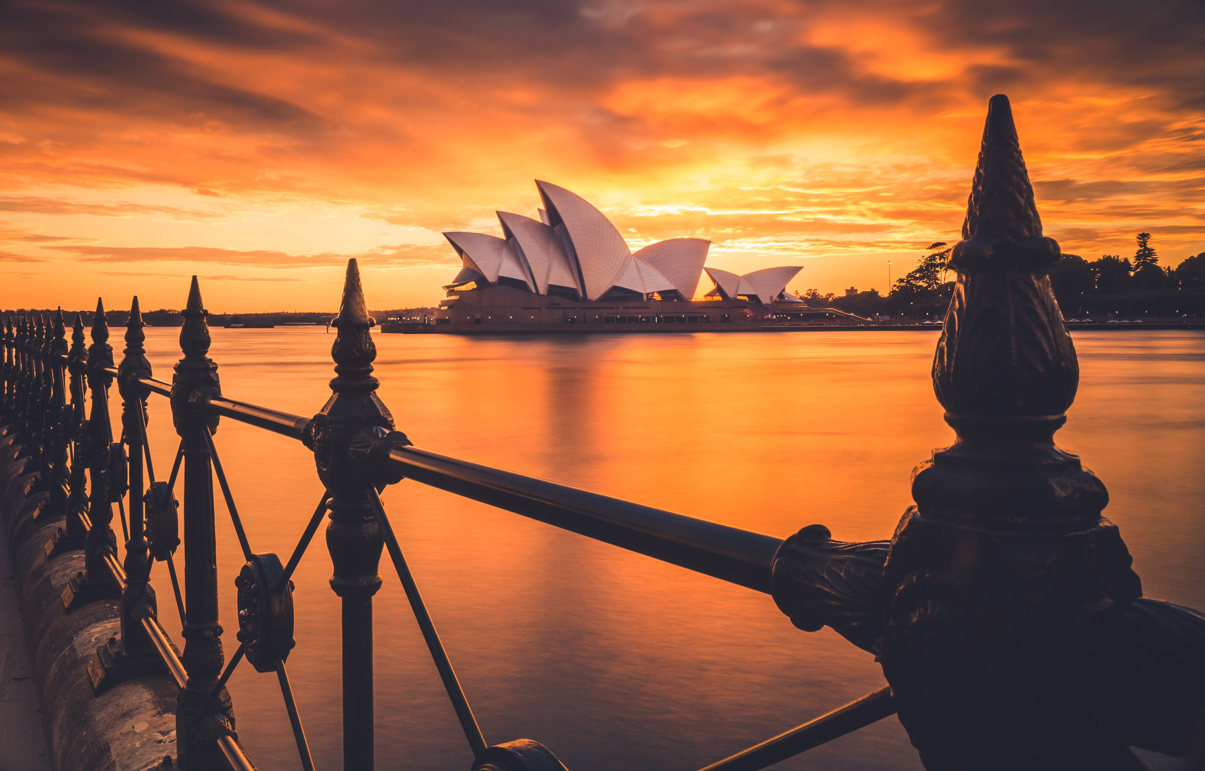 Sydney 桌布 壁紙 — 雪梨歌劇院在日落時閃耀金色光芒，背後有深色華麗欄杆，平靜水面反射火橙色雲彩。