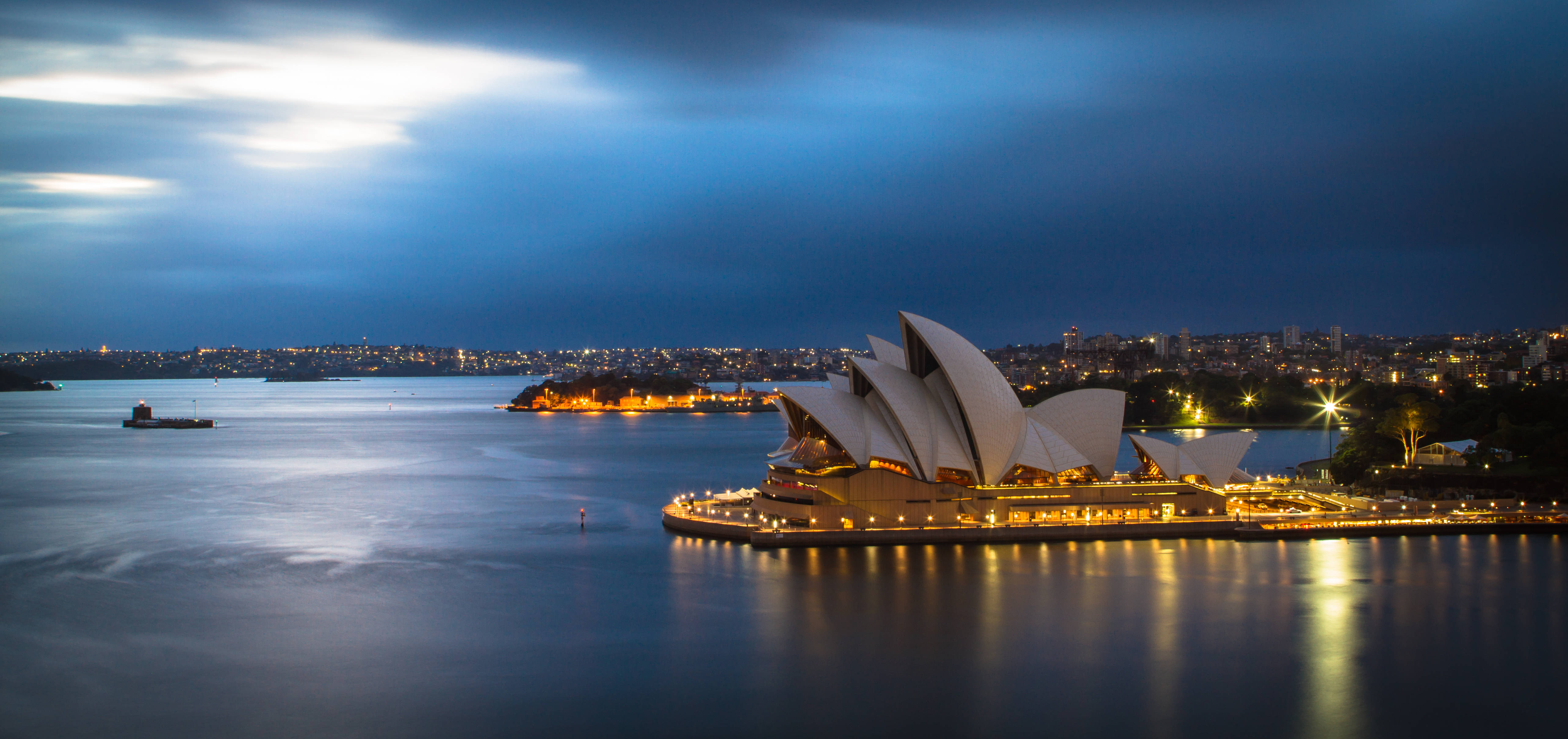 Sydney 桌布 壁紙 — 雪梨歌劇院在深藍色的暮色天空中溫暖地發光，其標誌性的帆船倒映在平靜的港灣水面上。