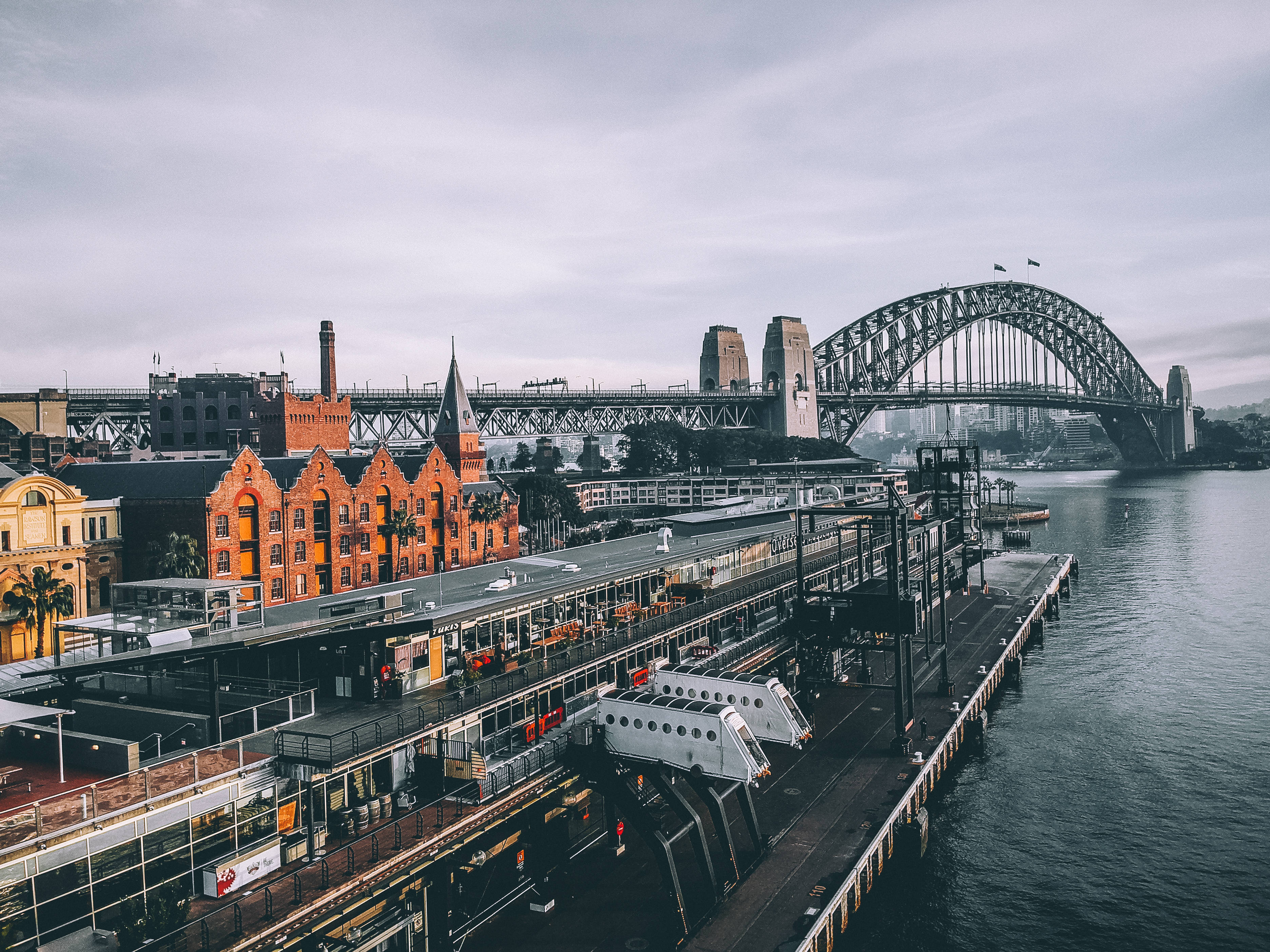 Sydney 桌布 壁紙 — 雪梨港灣大橋和歷史悠久的紅磚建築在平靜的水邊，陰天下的鳥瞰圖。